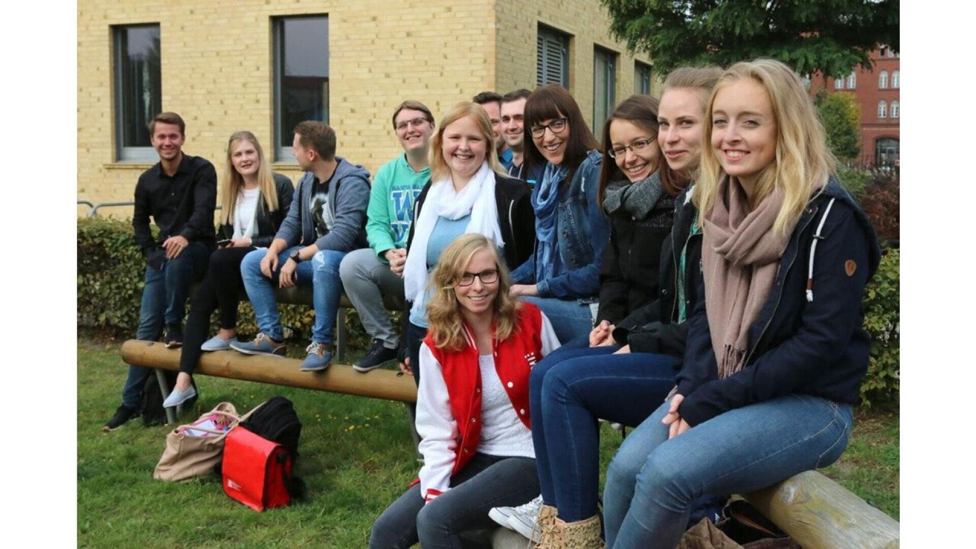 A group of smiling young adults sit together on wooden beams outdoors in front of a yellow brick building, posing for a group photo on a grassy area with bags placed nearby.