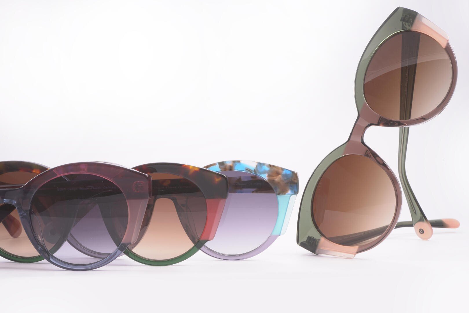 Vier stylische Sonnenbrillen mit bunten, durchscheinenden Rahmen sind vor weißem Hintergrund zu sehen. Drei sind nebeneinander aufgereiht, eine steht aufrecht dahinter.