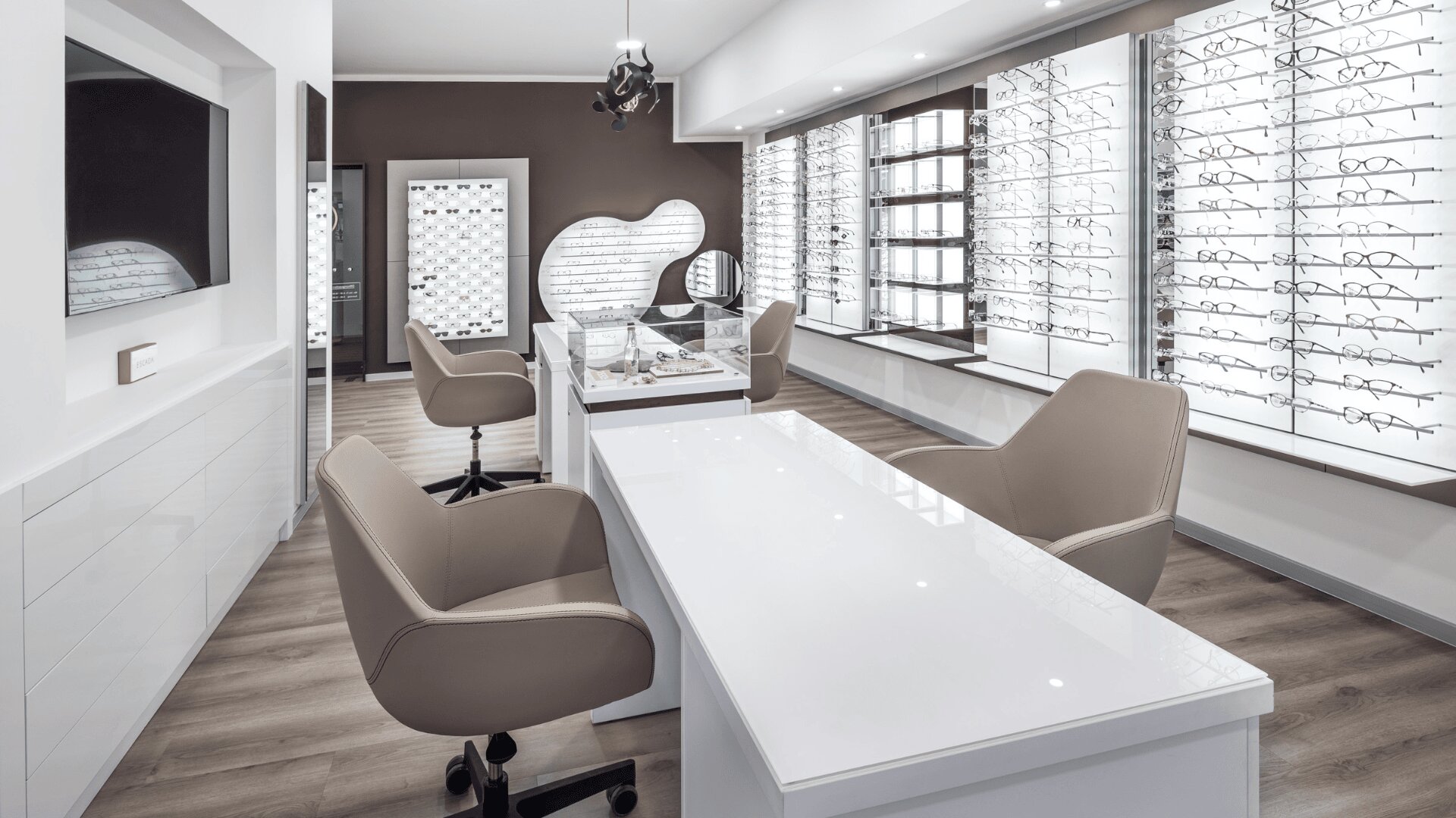 Modernes Optikerbüro mit beigen Stühlen, einem weißen Schreibtisch und Ausstellungswänden voller Brillengestelle. Der Raum hat einen Holzfußboden, helle Beleuchtung und ein sauberes, minimalistisches Design.