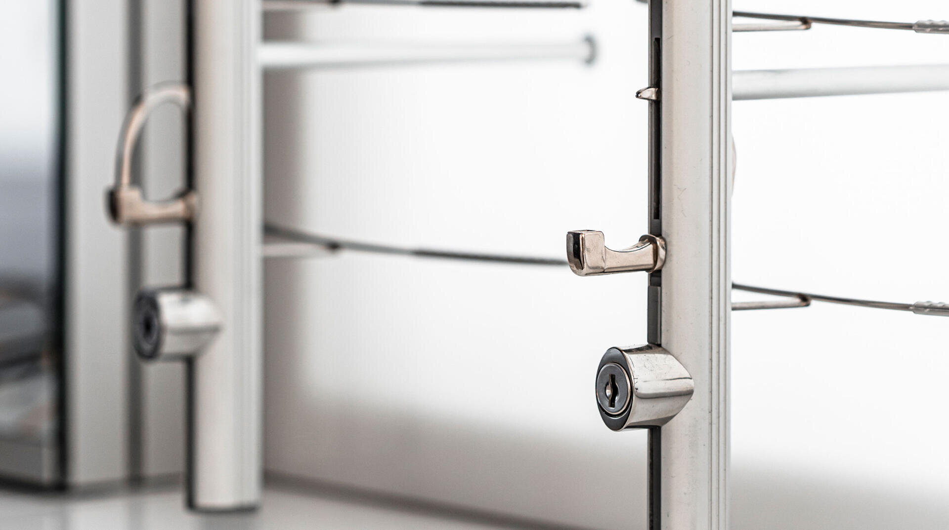 Nahaufnahme von zwei silbernen Metallschlössern, die vertikale Pfosten mit Kabeln sichern, vor einem weißen Hintergrund. Der Fokus liegt auf den Schlössern und hebt ihre Schlüssellöcher und Schließmechanismen hervor.