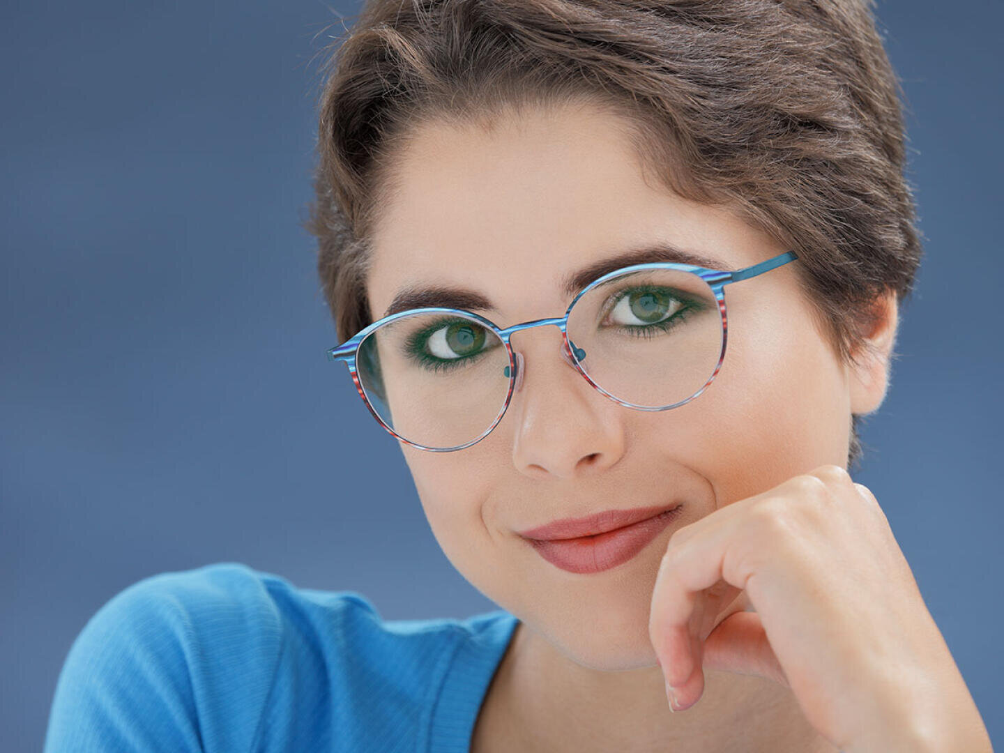A person with short brown hair, green eyes and blue round glasses smiles gently while resting her chin on her hand. She is wearing a blue top against a plain blue background.