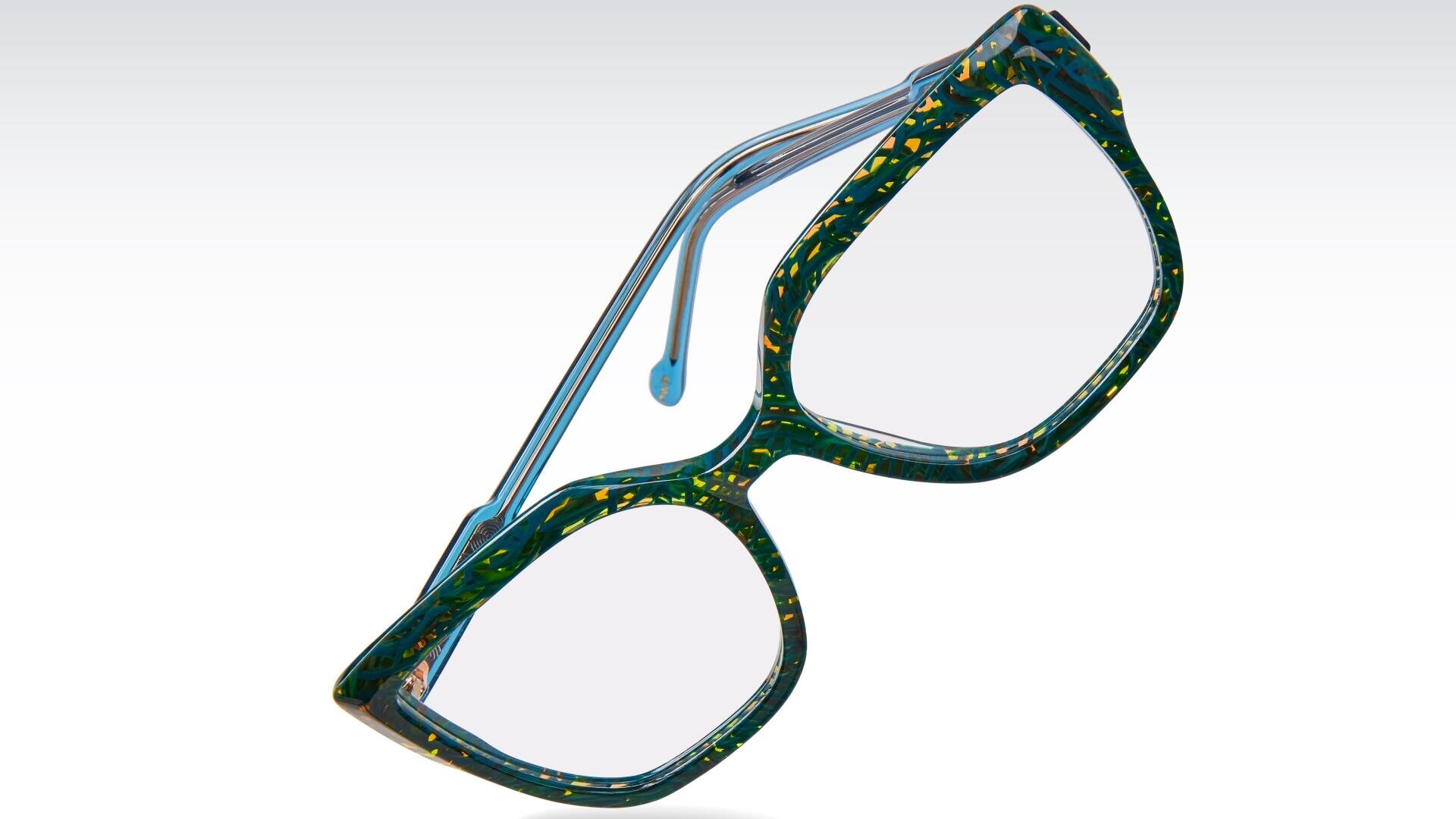 Eine dickrandige, geometrische Brille mit blau-grün marmoriertem Muster und gelben Sprenkeln auf weißem Hintergrund.