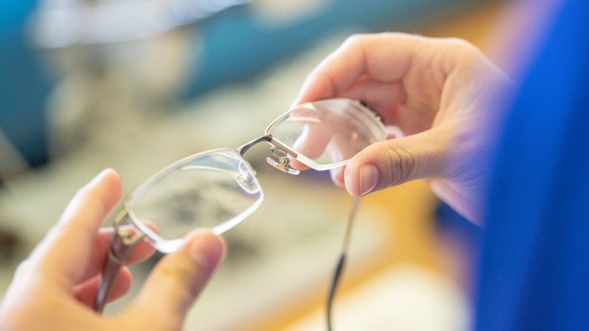 A person holds a pair of eyeglasses with both hands, inspecting the lenses closely. The background is blurred, drawing focus to the glasses and hands.