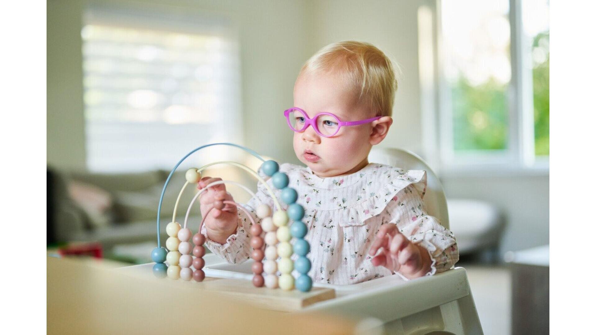 A baby wearing pink glasses sits at a table, playing with a wooden abacus toy with pastel-colored beads. The background shows a bright room with a window and a couch.