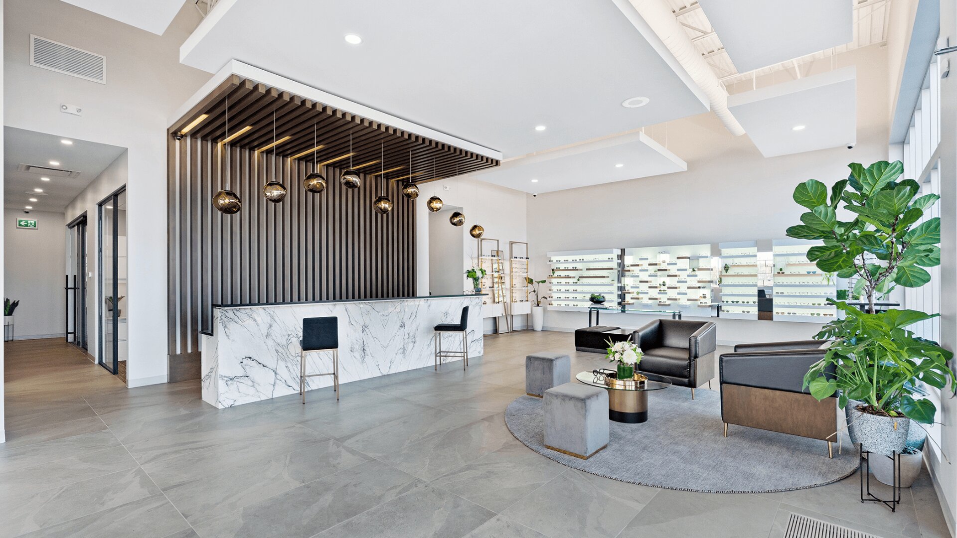 Modernes Foyer mit einer Empfangstheke aus Marmor, schwarzen Barhockern, goldenen Hängelampen, grauen Stühlen, kleinen Tischen, Zimmerpflanzen und großen Regalen an der Rückwand. Der Raum ist hell und minimalistisch gestaltet.