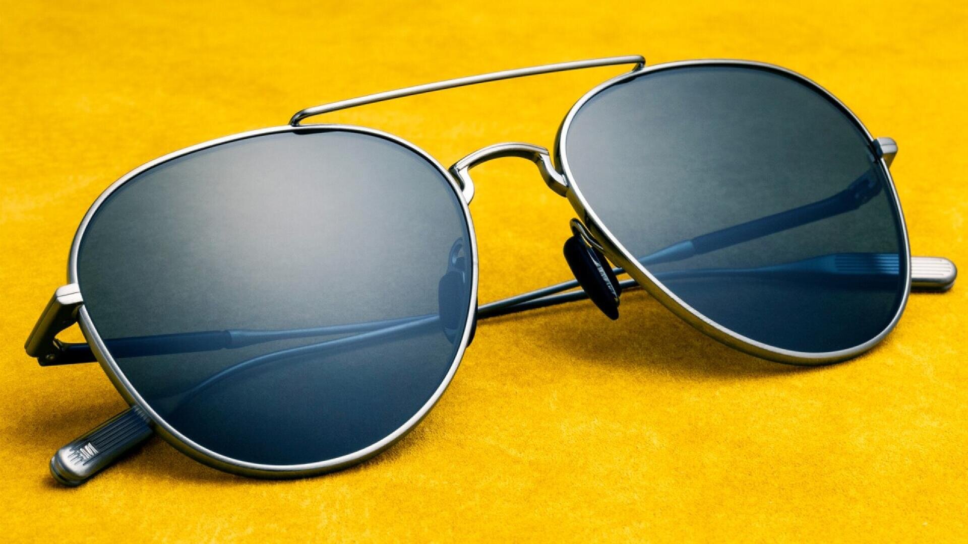 A pair of aviator-style sunglasses with dark lenses and a silver frame rests on a bright yellow surface.