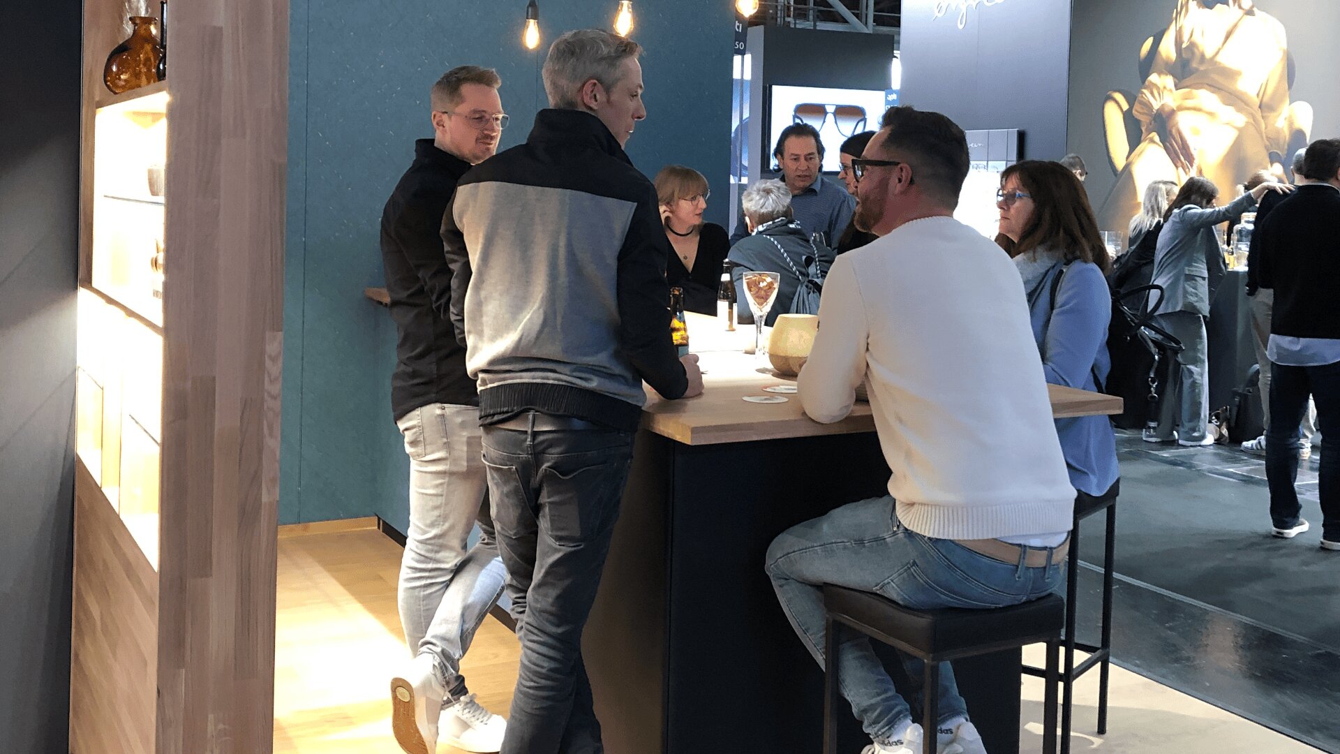 Eine Gruppe von Menschen sitzt und steht gemütlich um einen hohen Holztisch in einem modernen Innenraum und unterhält sich. Getränke und Gläser stehen auf dem Tisch, die Atmosphäre wirkt lebendig und gesellig.