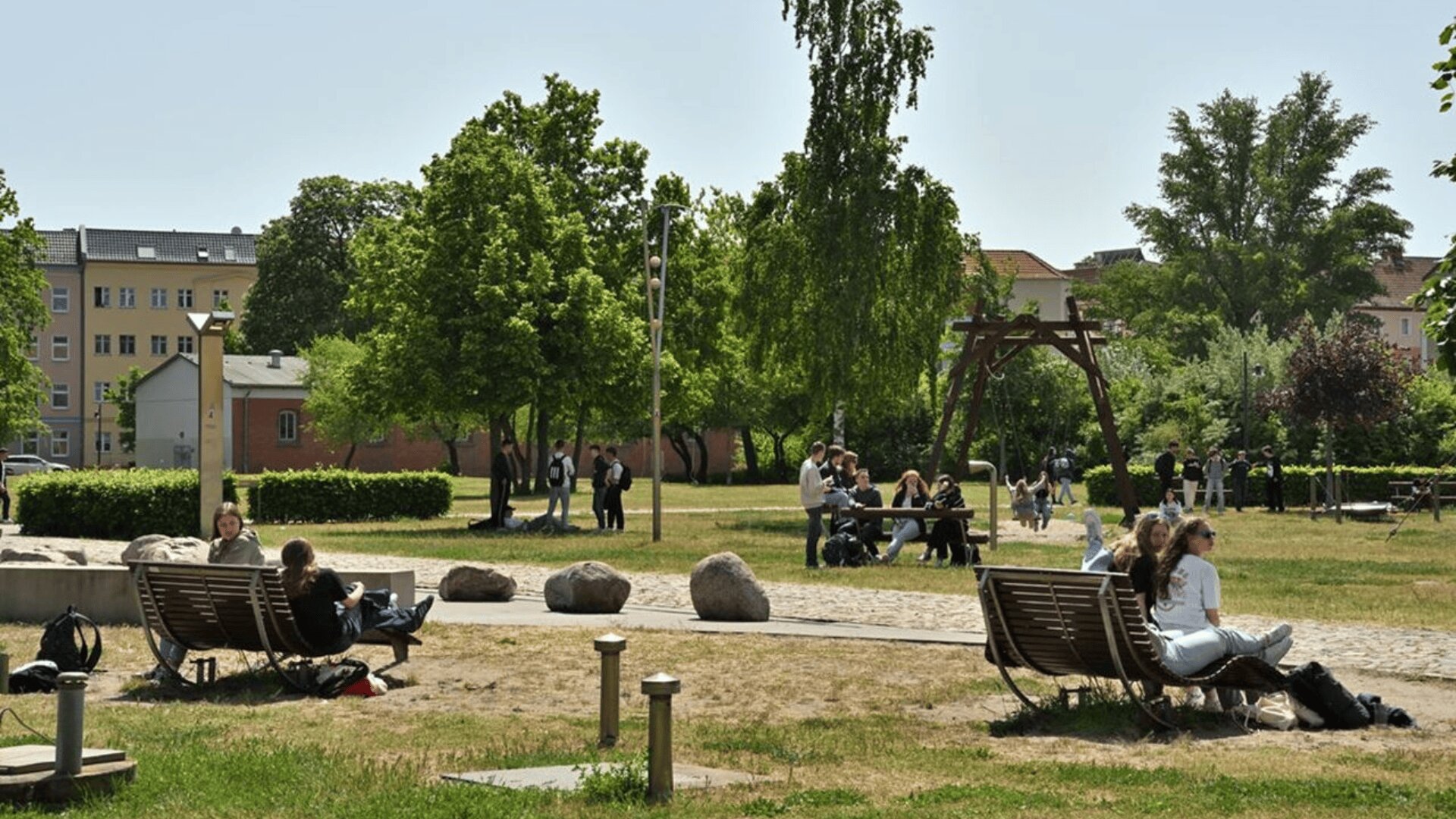 Menschen sitzen auf Bänken und spazieren in einem grünen Stadtpark mit Bäumen, Gras und einer Schaukel an einem sonnigen Tag herum. Im Hintergrund sind Gebäude zu sehen.
