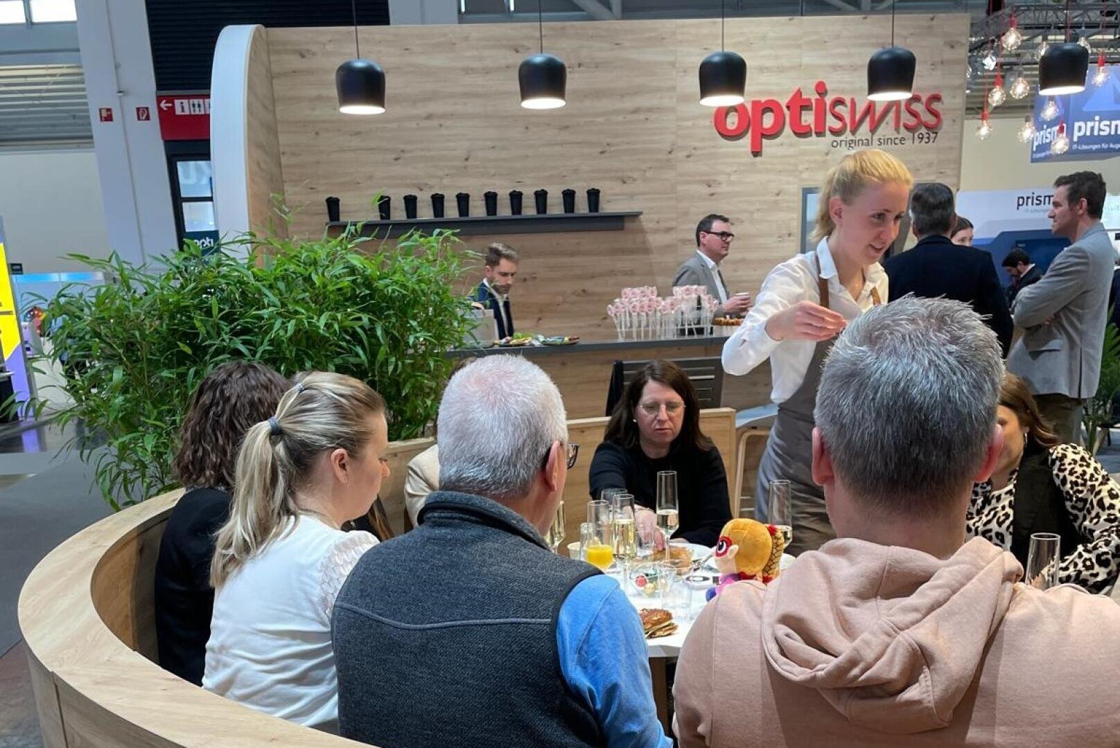 A group of people are seated around a table with food and drinks, while a server stands nearby. Behind them is a wooden booth with the optiswiss logo and shelves with cups, in a trade show or exhibition setting.