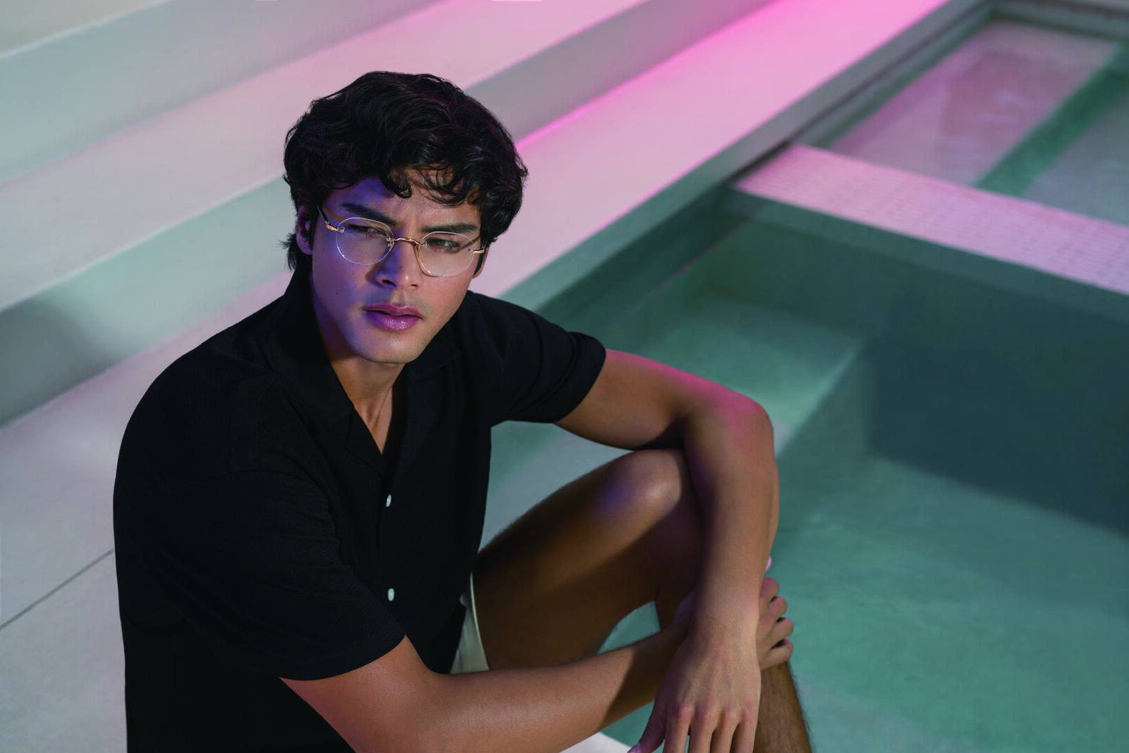 Eine Person mit Brille und schwarzem Hemd sitzt am Rande eines Swimmingpools mit Stufen, unter rosa und blauer Beleuchtung, und schaut nachdenklich.