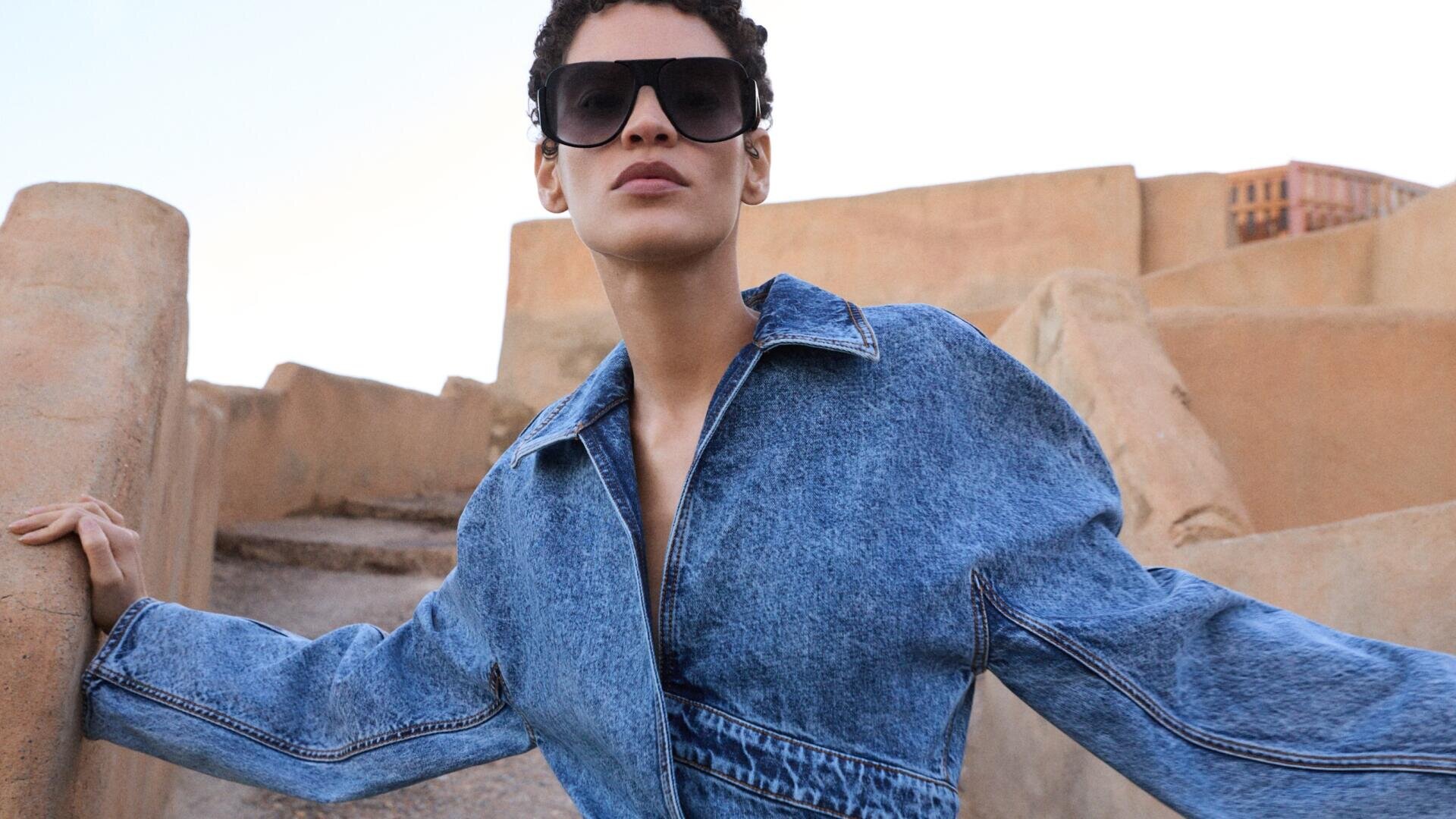 A person wearing a blue denim jacket and large dark sunglasses poses outdoors against tan stone steps and walls, with a clear sky in the background.