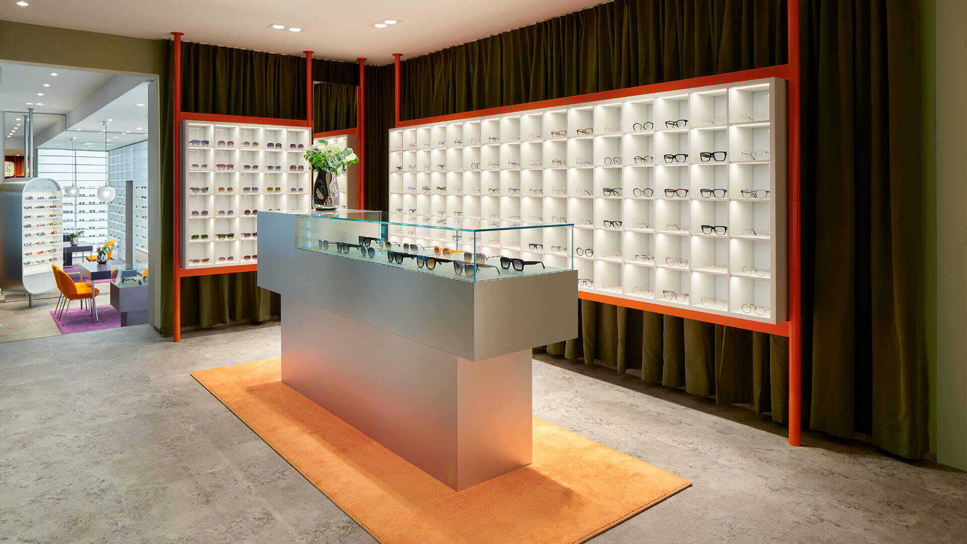Modernes Brillengeschäft mit einer Glasvitrine auf einem orangefarbenen Teppich, in der verschiedene Brillen ausgestellt sind. Dahinter hängen Regale mit Brillengestellen an einer Wand mit dunkelgrünen Vorhängen. Das Geschäft ist hell erleuchtet.