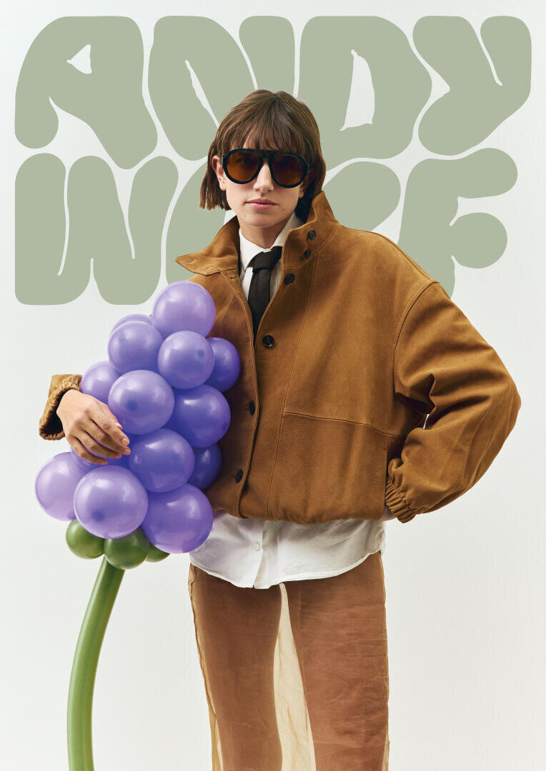 A person wearing sunglasses, a brown jacket and a white shirt is holding a bunch of purple balloon grapes. An abstract green text with the inscription ANDY WOLF can be seen in the background.