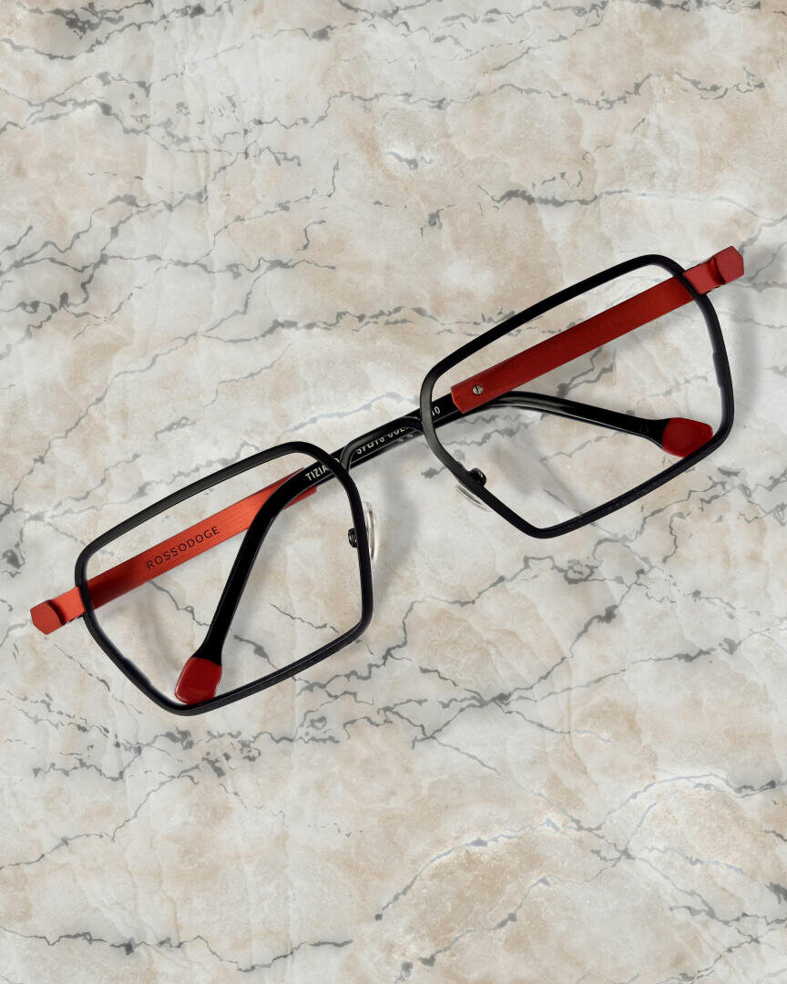 A rectangular pair of glasses with a black frame and red temples rests on a light marble surface with gray and light brown veining.