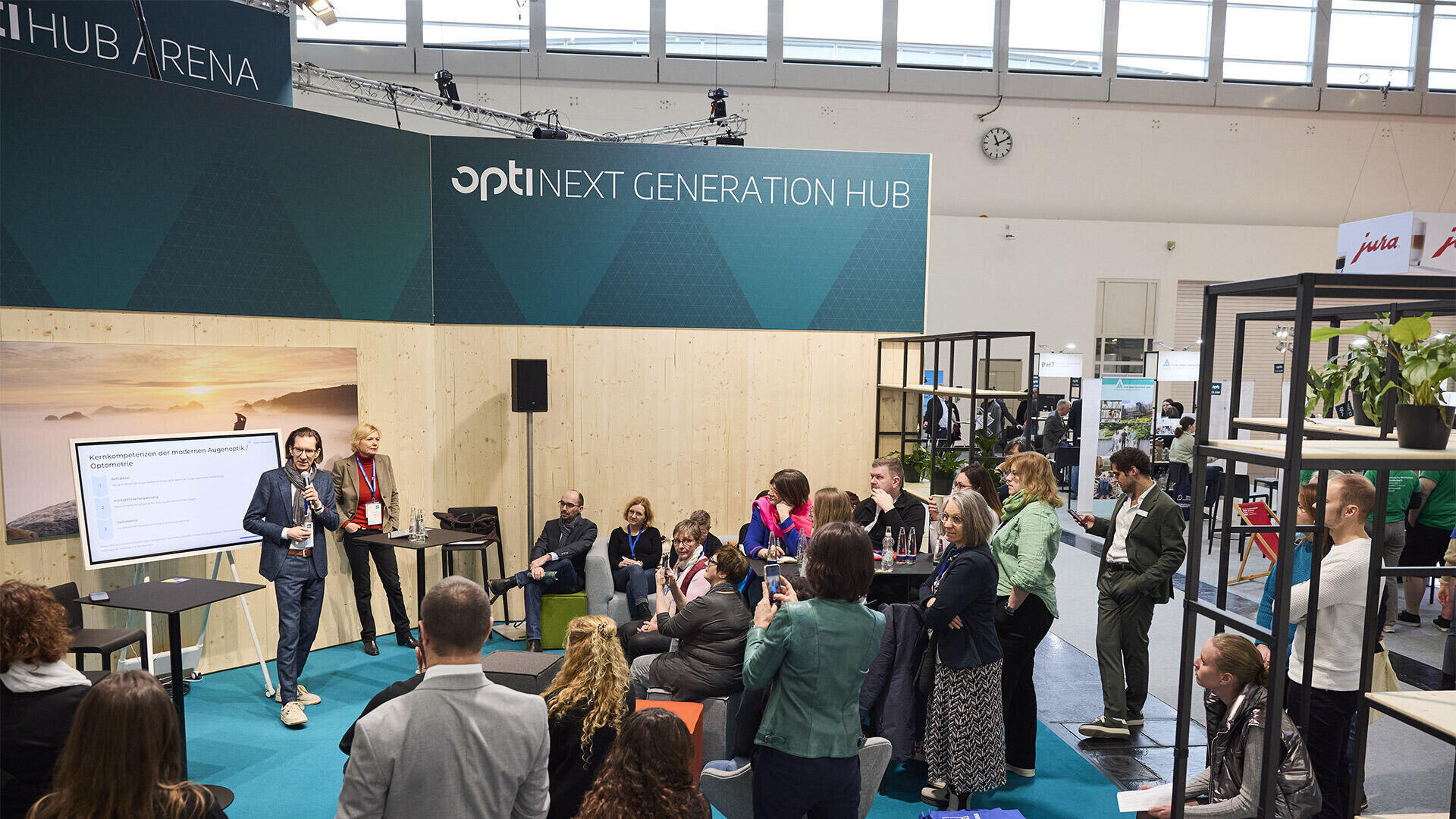 A speaker gives a presentation to an attentive audience at a conference in a modern, open room with a screen and signage that reads Next Generation Hub. People are sitting and standing, some taking notes or photos.
