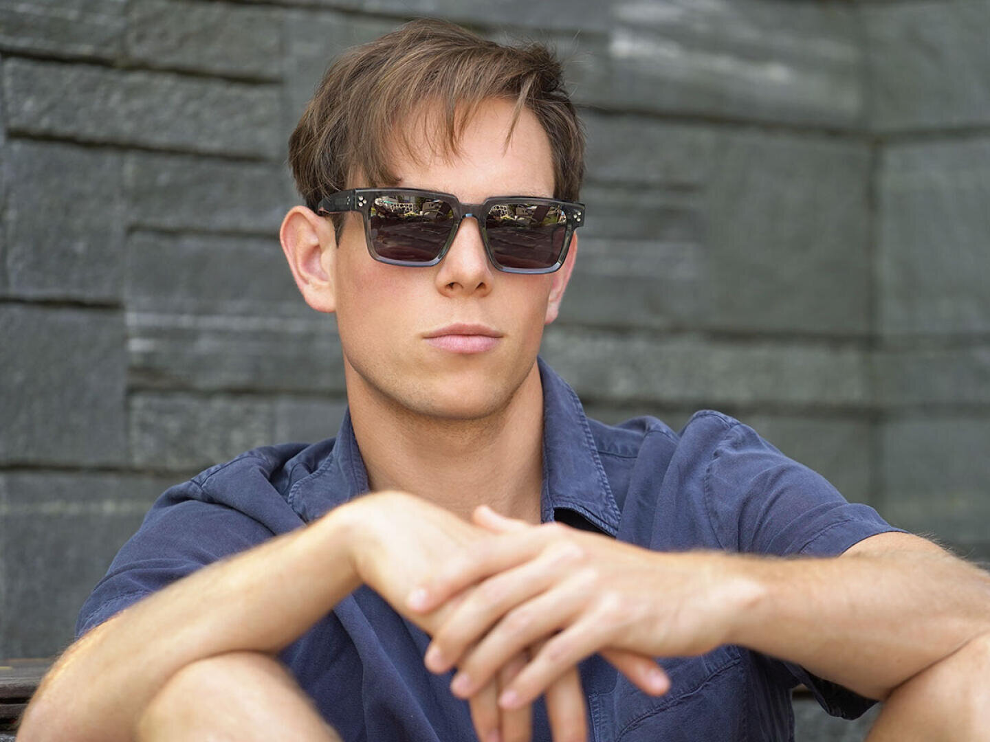A young man with short brown hair, dark sunglasses and a blue shirt is sitting outside against a stone wall, resting his arms on his knees and looking slightly to the side with a neutral expression.