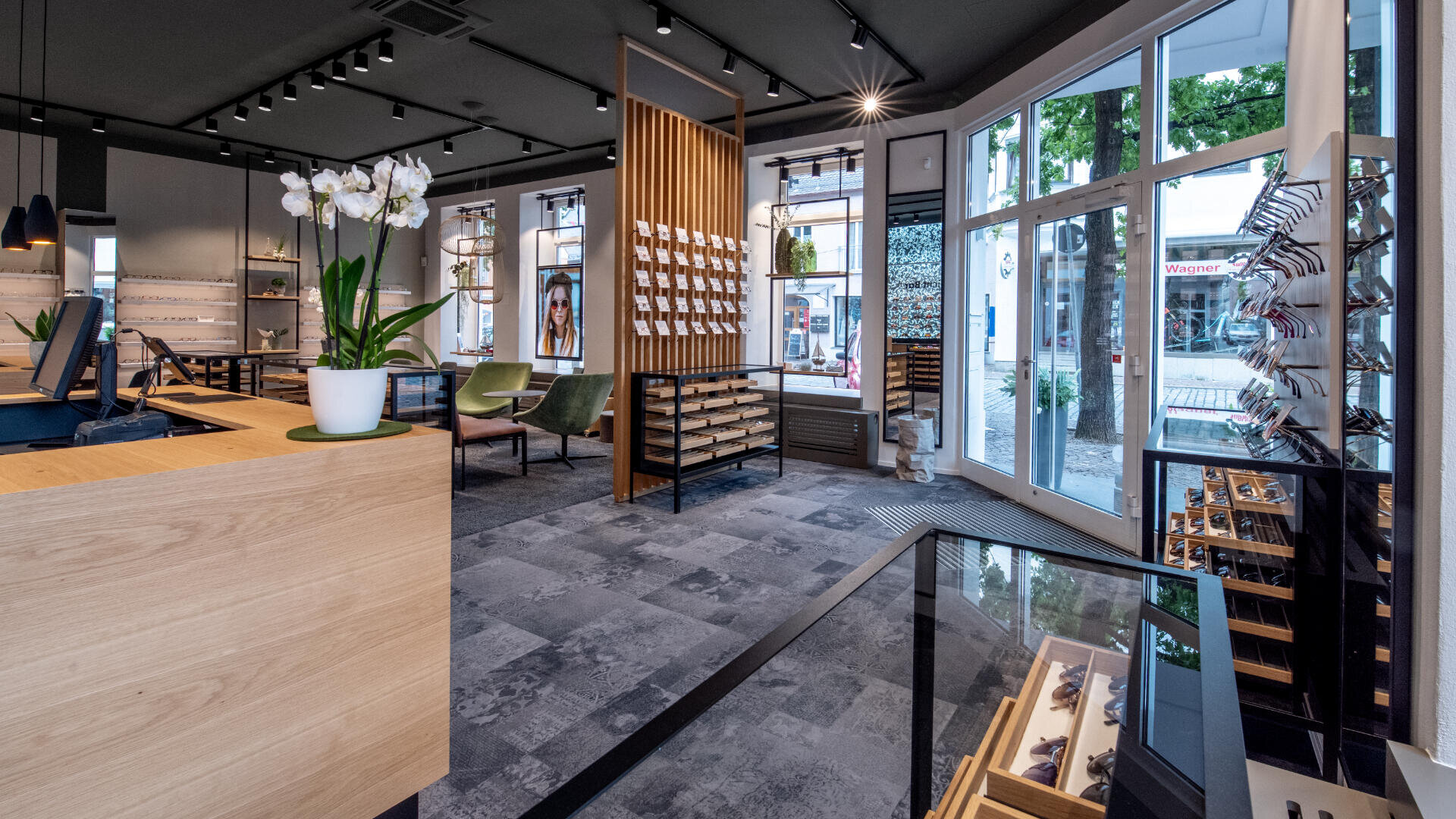 Modernes Interieur eines Optikergeschäfts mit Brillenregalen, einer Holztheke mit einer Orchidee im Topf, einem Sitzbereich mit grünen Stühlen, großen Fenstern an der Vorderseite und viel natürlichem Licht.