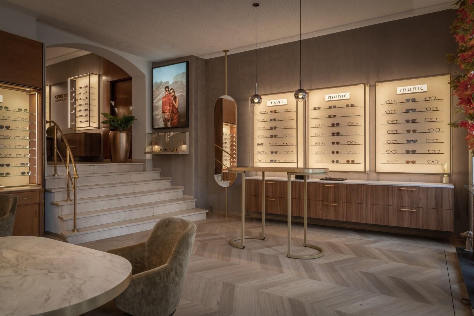 Ein stilvolles Optikergeschäft mit Brillen auf beleuchteten Wandregalen, einem Marmortisch mit Samtstühlen, einer Treppe mit goldenem Handlauf und einem großen Spiegel an der Wand. Warmes, modernes Dekor und sanfte Beleuchtung.
