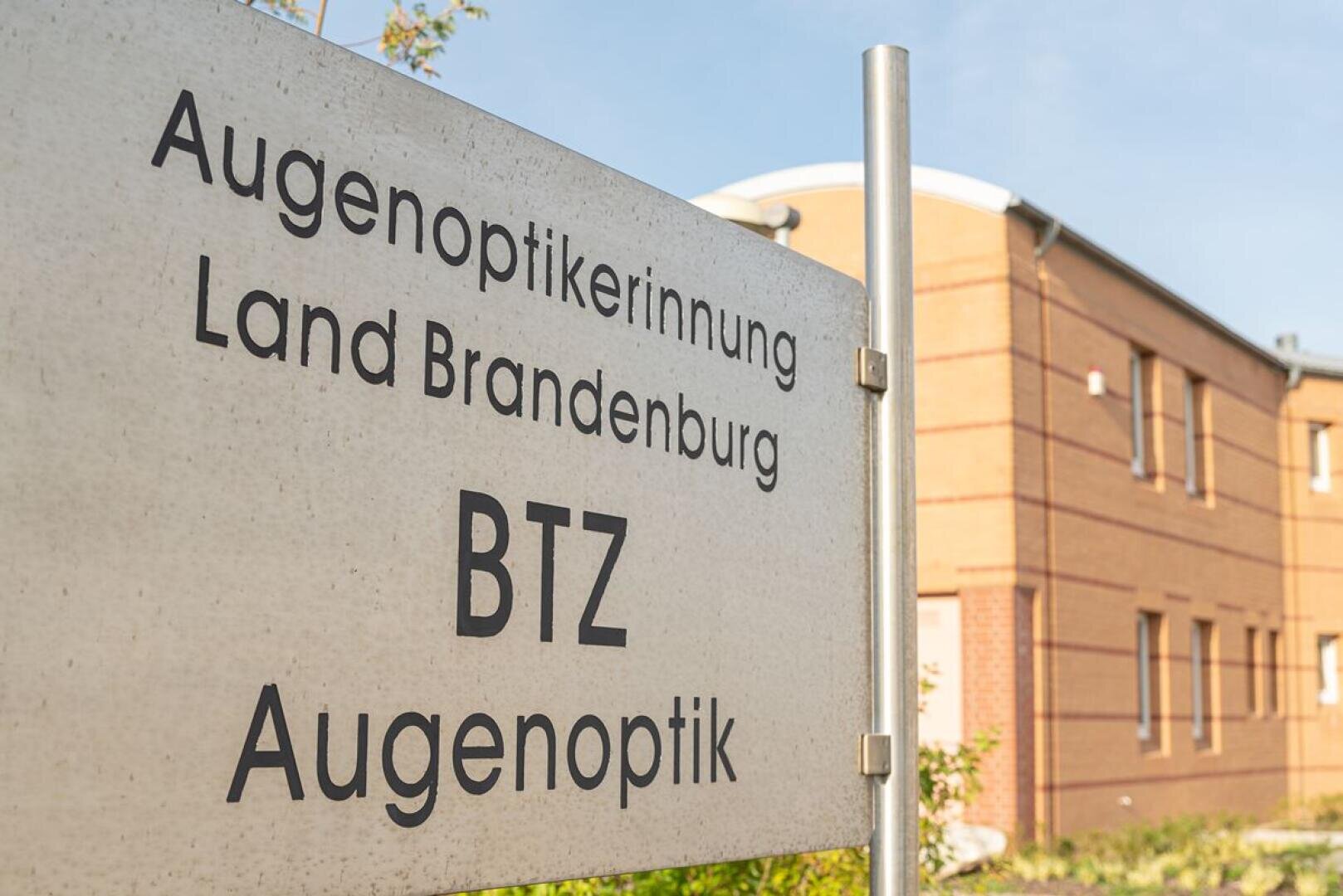 A sign reading "Augenoptikerinnung Land Brandenburg BTZ Augenoptik" stands in front of a modern brick building on a sunny day.