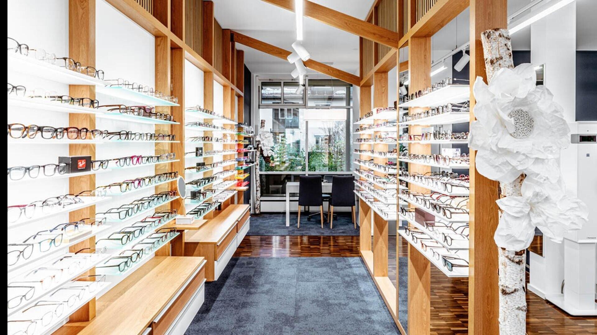 A modern eyewear store interior with wooden beams, white shelves displaying many pairs of eyeglasses, and a seating area by a large front window. A decorative white flower is attached to a birch branch on the right.