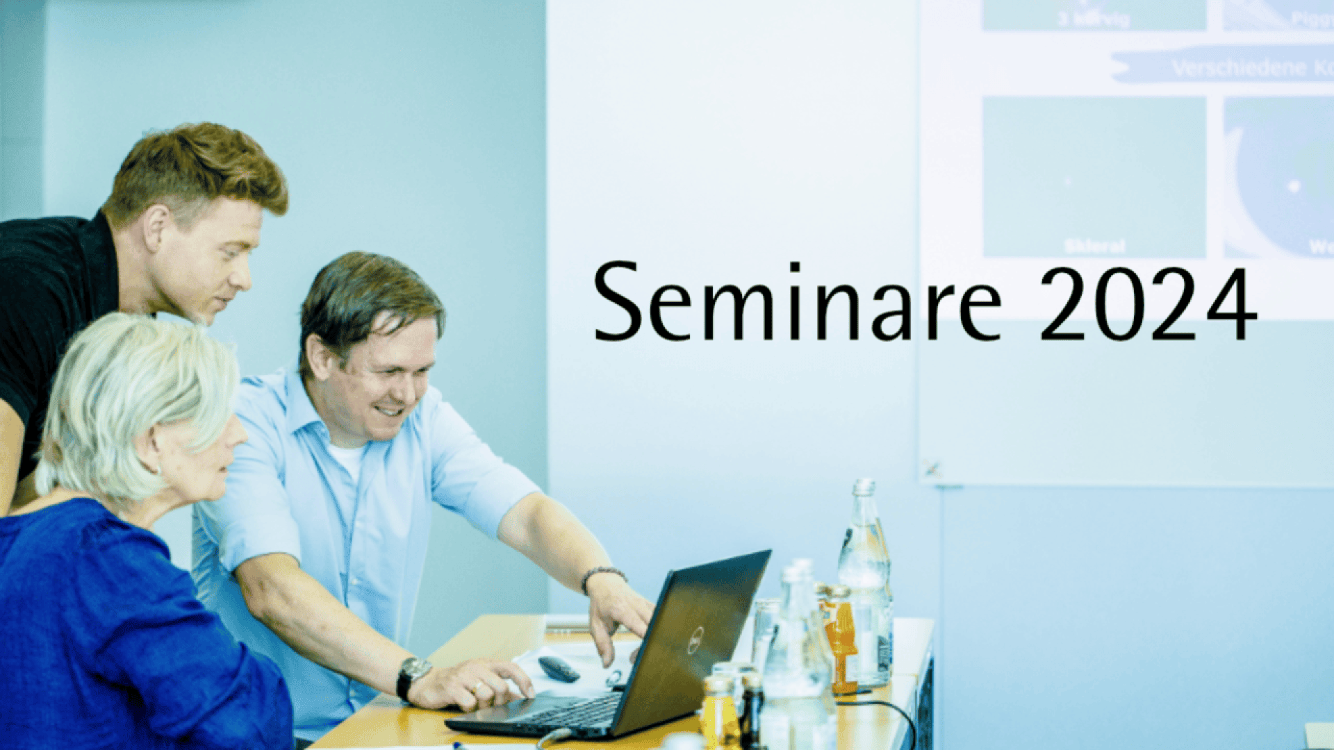 Three people sit and stand around a laptop, engaged in discussion in a bright room. The words Seminare 2024 are displayed on the wall beside them. Bottles and glasses are on the table.