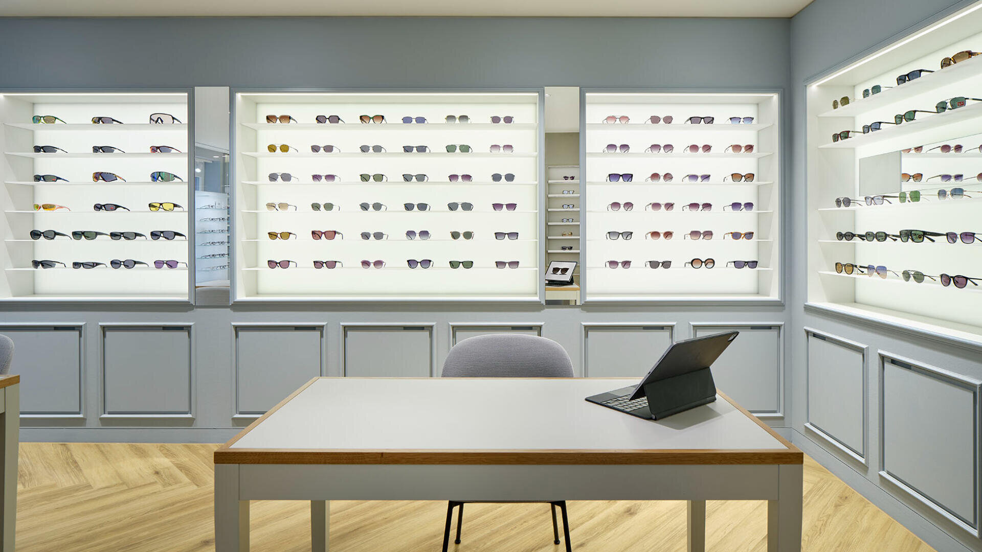 Die Inneneinrichtung eines modernen Brillengeschäfts ist mit Reihen von Brillen und Sonnenbrillen auf beleuchteten Regalen ausgestattet; im Vordergrund steht auf einem Holzboden ein Tisch mit einem Stuhl und einem Tablet.