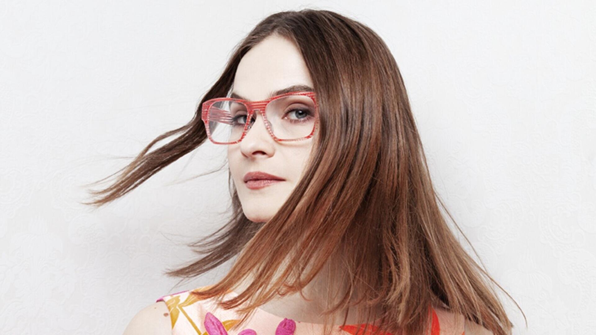 A woman with straight brown hair wearing pink glasses and a floral top looks at the camera, her hair swept to the side against a plain white background.