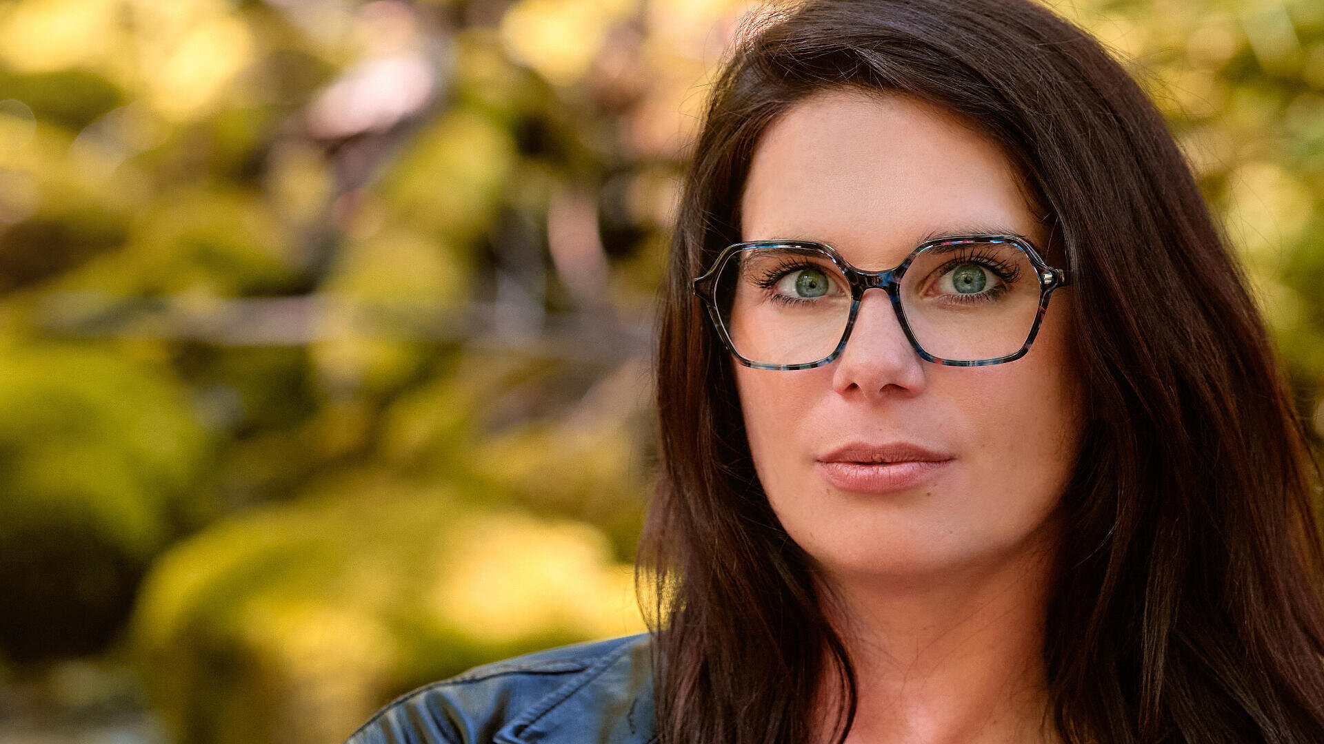 Eine Frau mit langen braunen Haaren und großer Brille blickt in die Kamera. Sie ist im Freien vor einem unscharfen Hintergrund aus grünem und gelbem Laub.