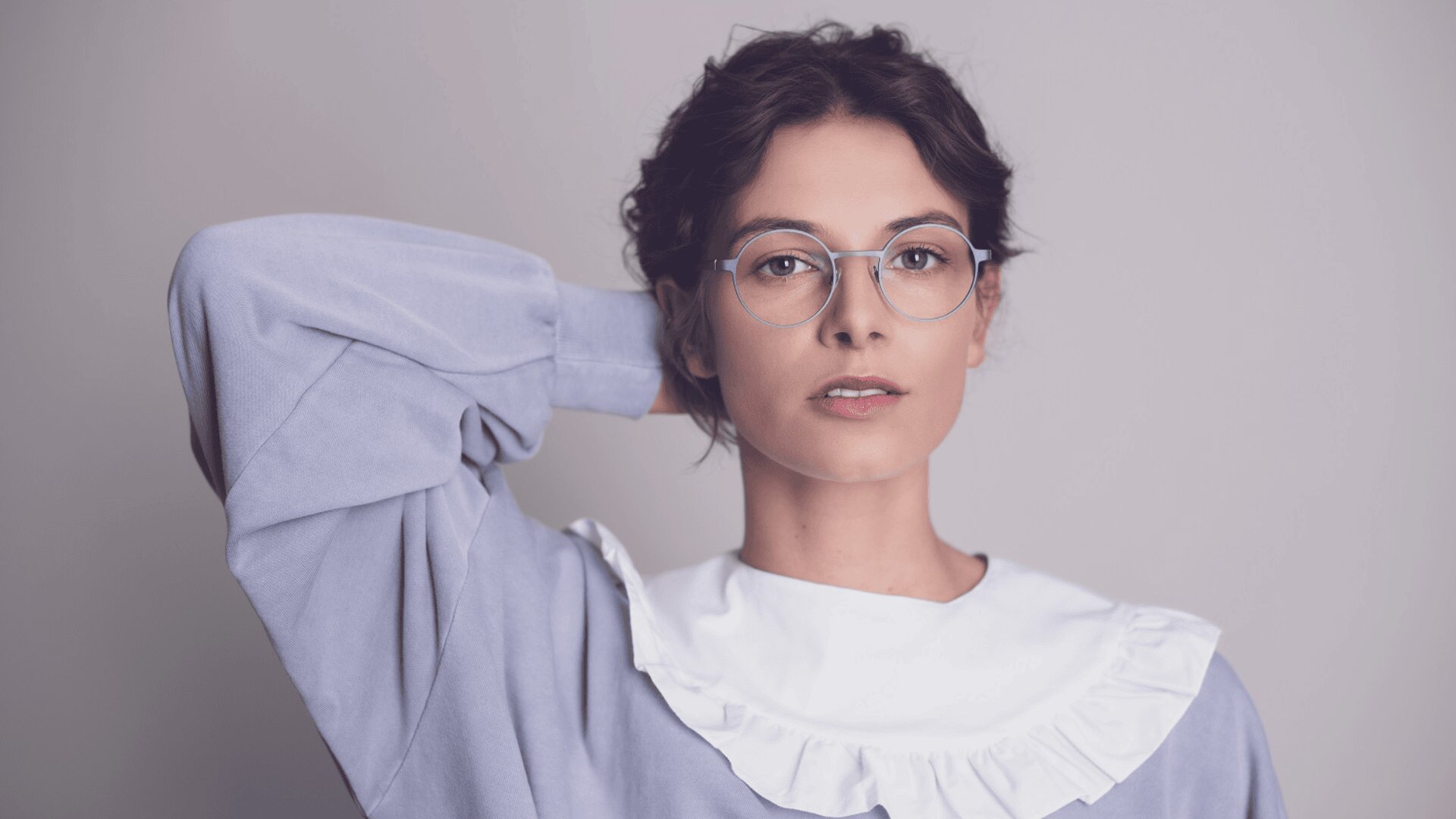 A woman with short, wavy dark hair wears round glasses and a light purple top with a white ruffled collar, posing with one hand resting behind her head against a plain background.