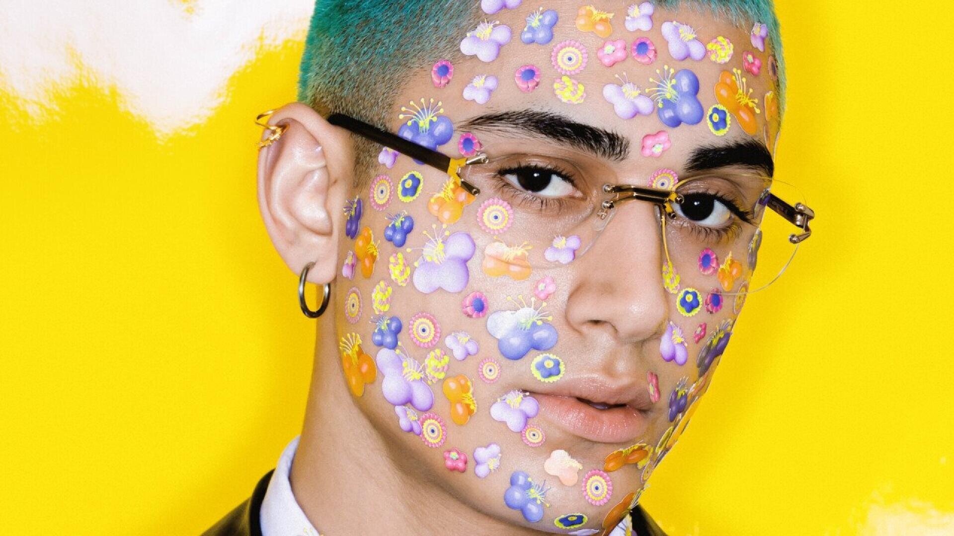 A person with teal hair and glasses poses against a bright yellow background. Their face is covered in colorful stickers shaped like flowers, butterflies, and smiley faces. They wear a small hoop earring and a calm expression.