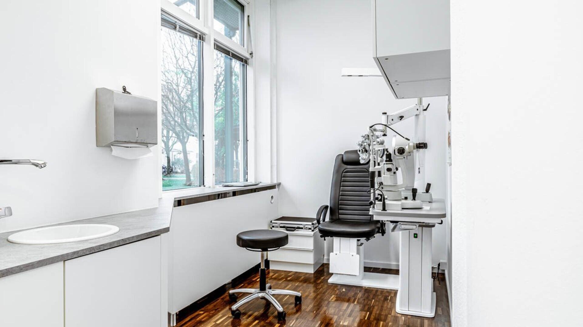 A clean, modern eye examination room with an exam chair, eye testing equipment, a stool, a sink, a paper towel dispenser, and large windows letting in natural light.
