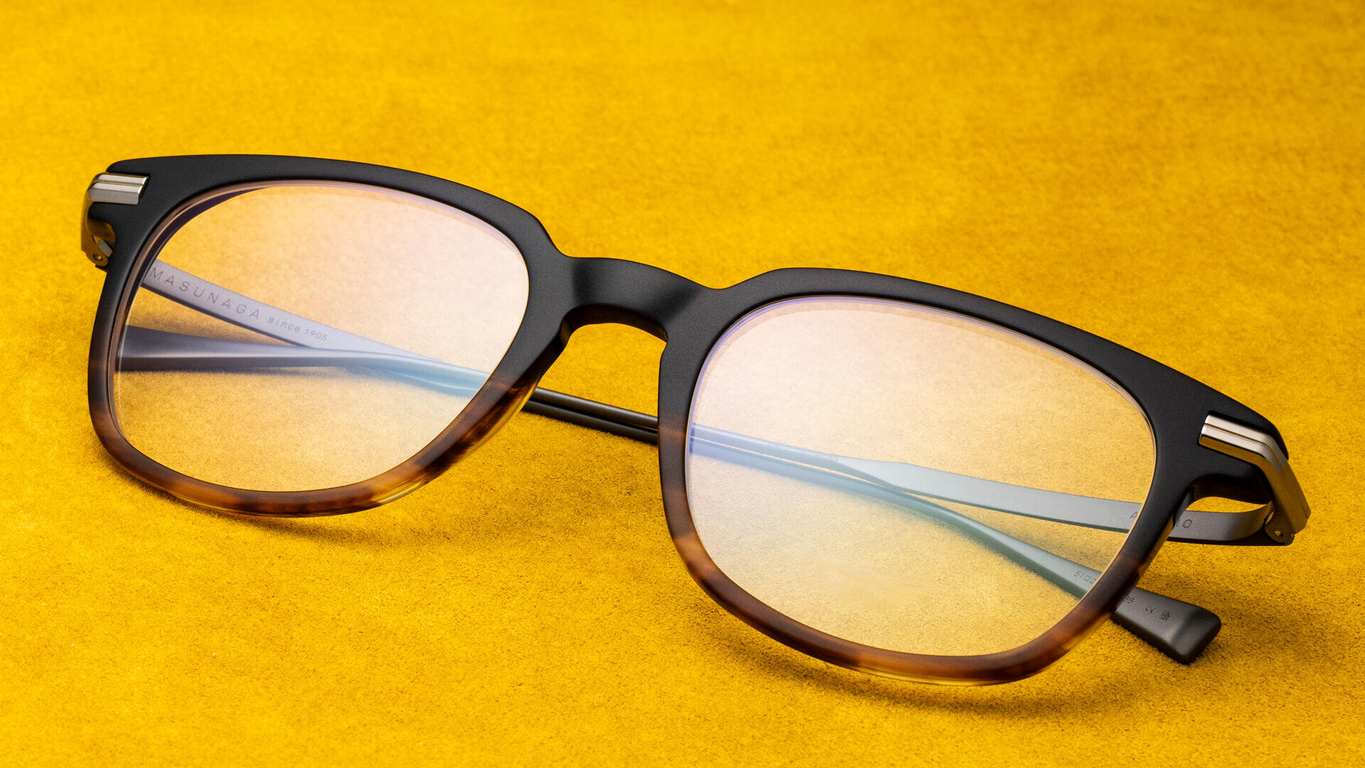 Eine stilvolle Brille mit schwarz-braunem Schildpattrahmen liegt auf einer leuchtend gelben Oberfläche. Die Brille hat leicht getönte Gläser und Metallakzente an den Ecken.