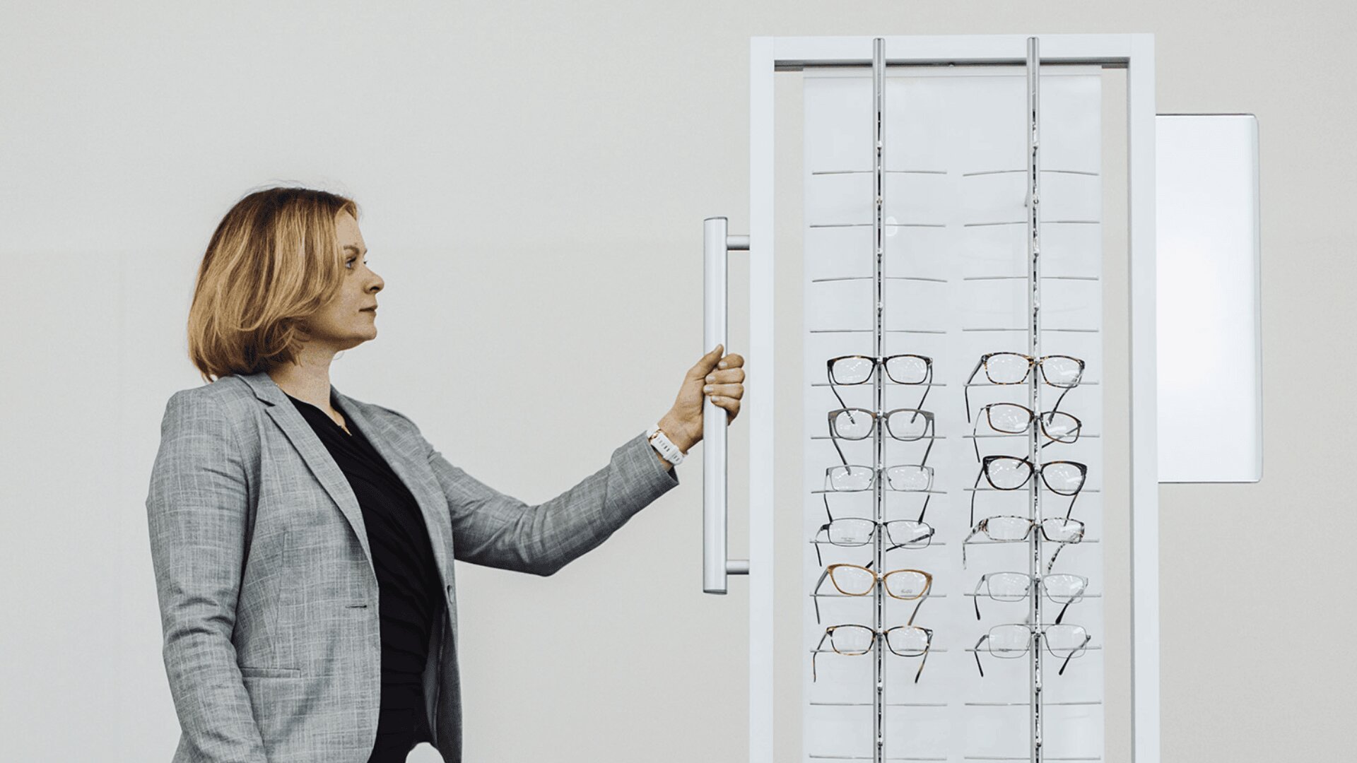 A woman wearing a gray blazer stands next to a display rack filled with various eyeglasses, looking at the frames and holding the handle of the display.