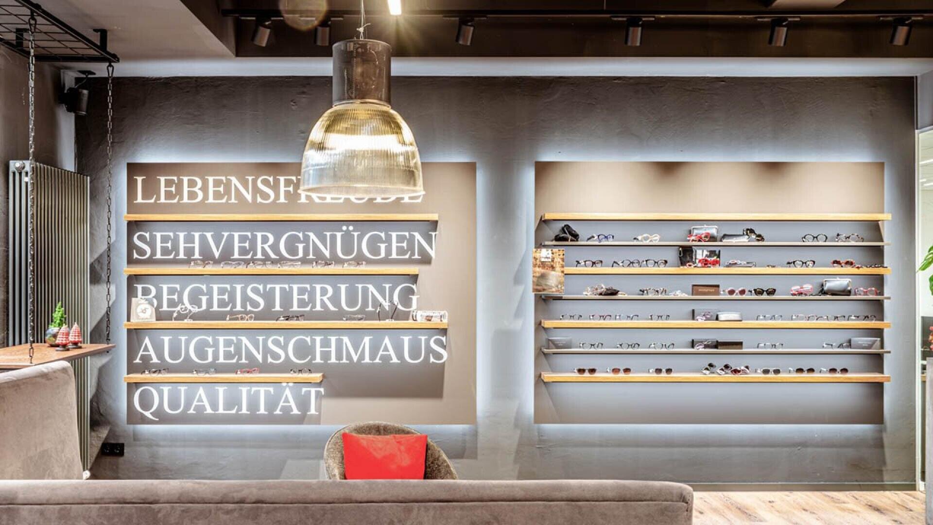 Modern eyewear store interior with stylish lighting, a gray couch, and illuminated German words on the wall. Shelves on the right display various eyeglasses and sunglasses against a gray background.