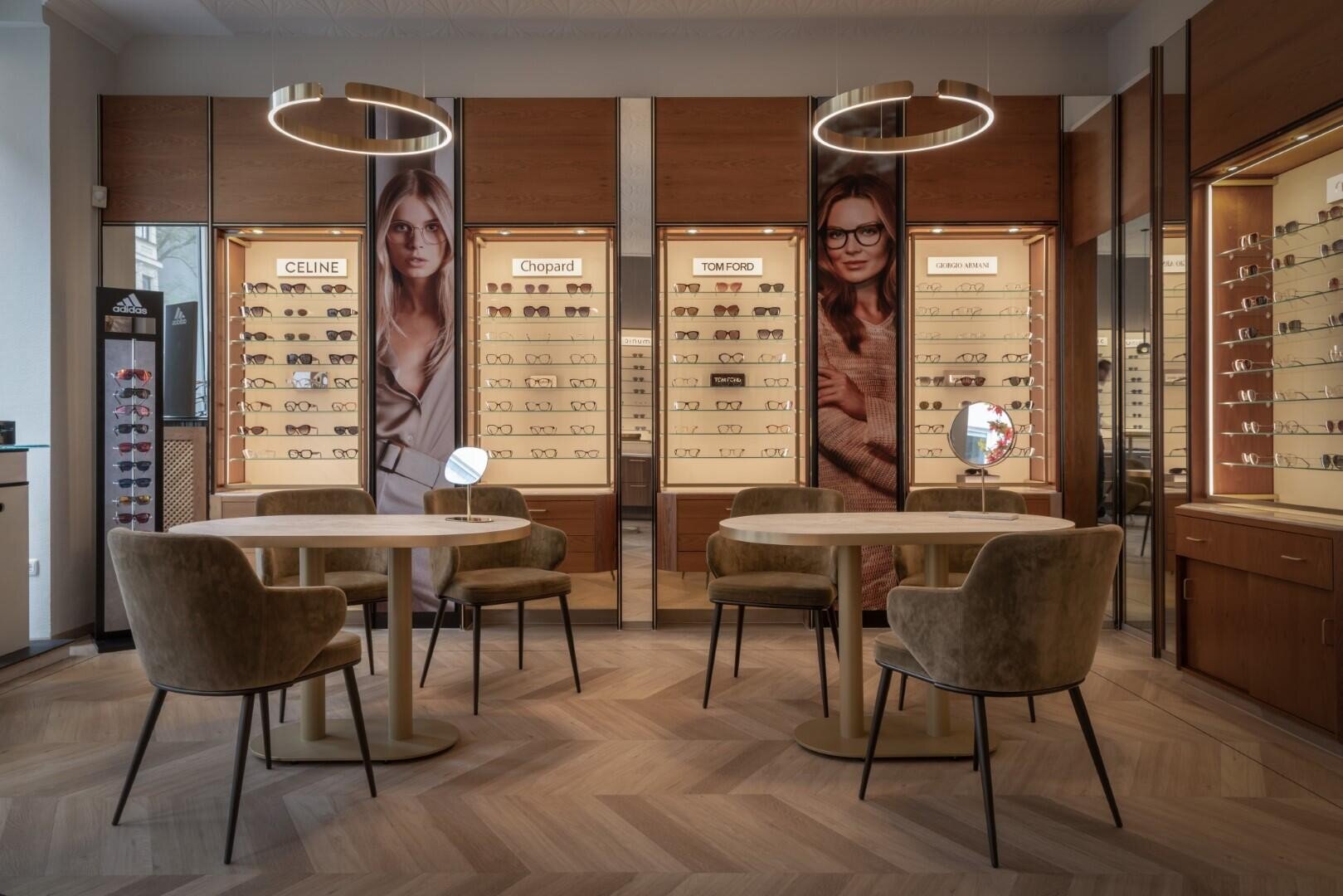 Ein modernes Brillengeschäft mit Holzregalen, stilvollen runden Tischen und Stühlen sowie großen Fotos von Models mit Brillen an den Wänden. Warmes Licht schafft eine einladende Atmosphäre.