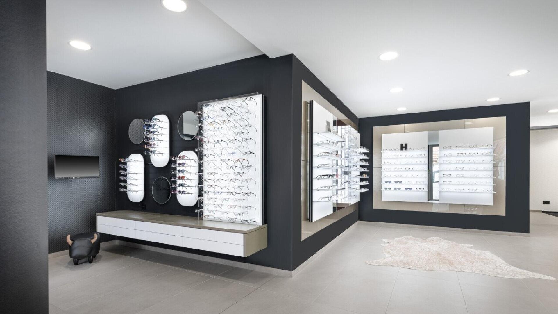 A modern optician’s store interior with sleek display walls featuring numerous eyeglasses, several round mirrors, a wall-mounted TV, and minimalist white and gray decor. A small rug and a decorative stool are on the floor.