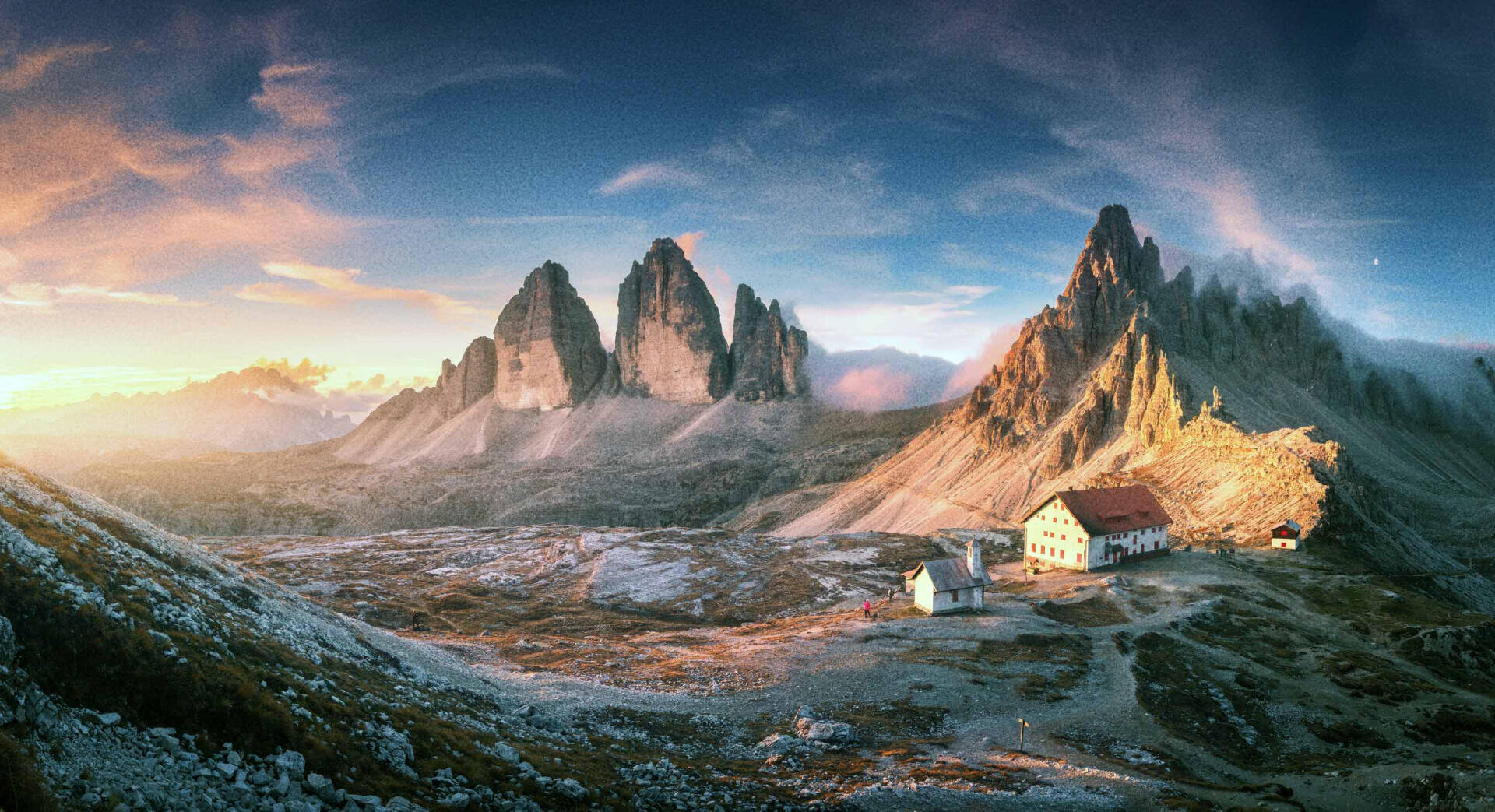 Ein Panoramablick auf zerklüftete Berggipfel bei Sonnenaufgang, wobei das Sonnenlicht eine Ansammlung von Gebäuden in einer felsigen Alpenlandschaft beleuchtet. Der Himmel ist teilweise bewölkt und wirft dramatische Schatten auf die Szene.