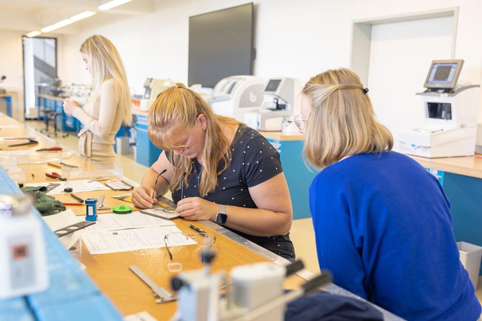 Three women are working in a bright laboratory; two of them are concentrating on a practical project at a desk with tools and papers, while another stands in the background operating a telephone or device. Lab equipment can be seen around them.