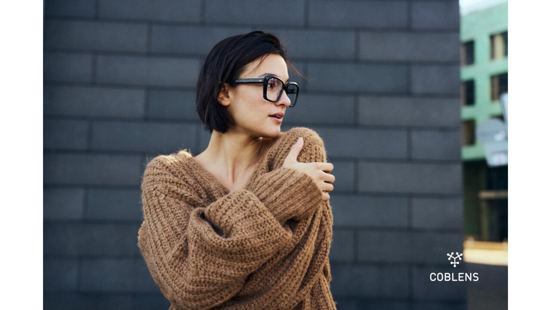 A woman with short dark hair wearing large black glasses and a chunky brown sweater stands in front of a dark brick wall, with “COBLENS” and a small logo in the bottom right corner.