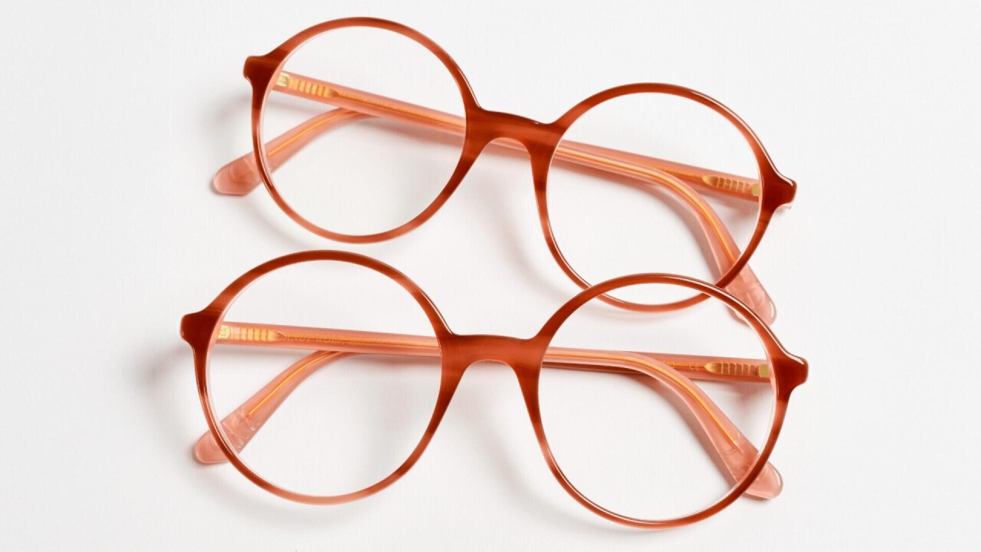Two pairs of round, orange eyeglasses are stacked on a white surface, with the top pair slightly overlapping the bottom pair.