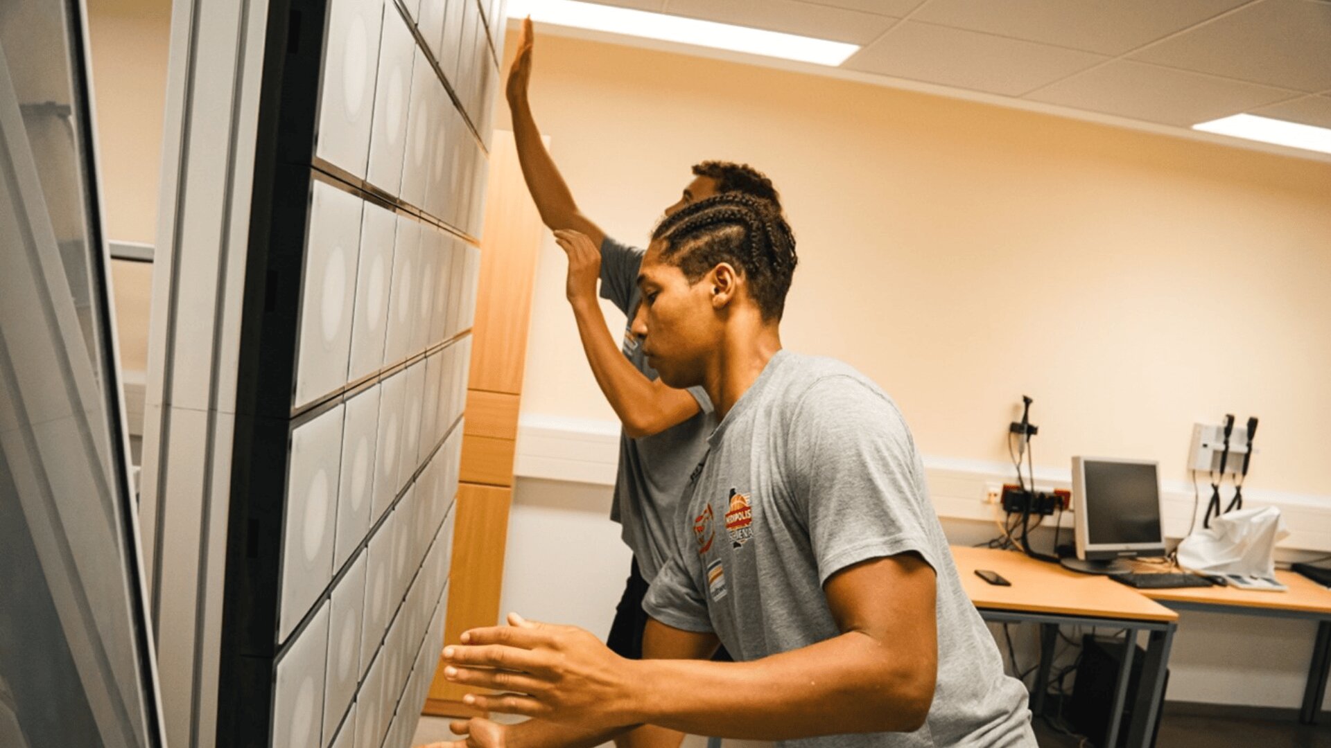 Zwei Personen in grauen Hemden stehen in einem Raum mit Schreibtischen und Computern. Sie blicken auf eine große vertikale Tafel mit mehreren quadratischen Ausschnitten und interagieren mit ihr.