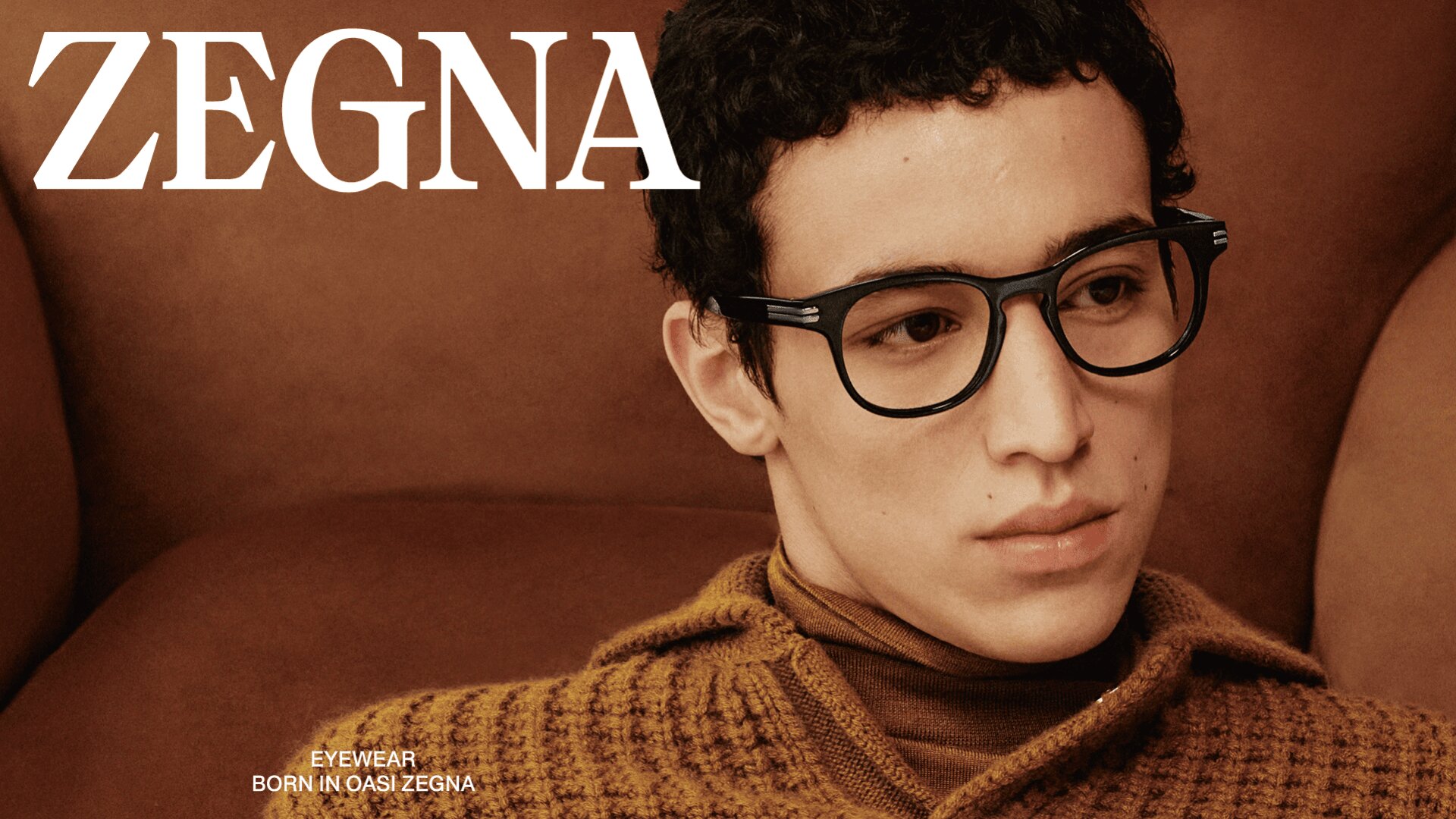 Ein junger Mann mit einer schwarzen Brille und einem braunen Strickpullover sitzt auf einem braunen Stuhl. Oben links steht in fetten weißen Buchstaben das Wort ZEGNA, darunter in kleinerer Schrift: EYEWEAR BORN IN OASI ZEGNA.