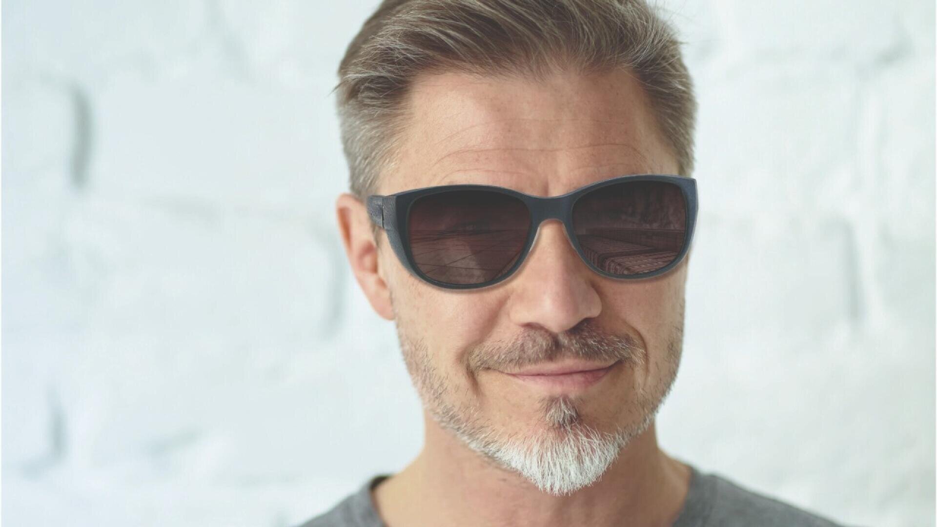 A middle-aged man with short brown hair, a salt-and-pepper beard, and mustache smiles slightly while wearing black sunglasses and a gray shirt, standing in front of a light-colored, textured wall.