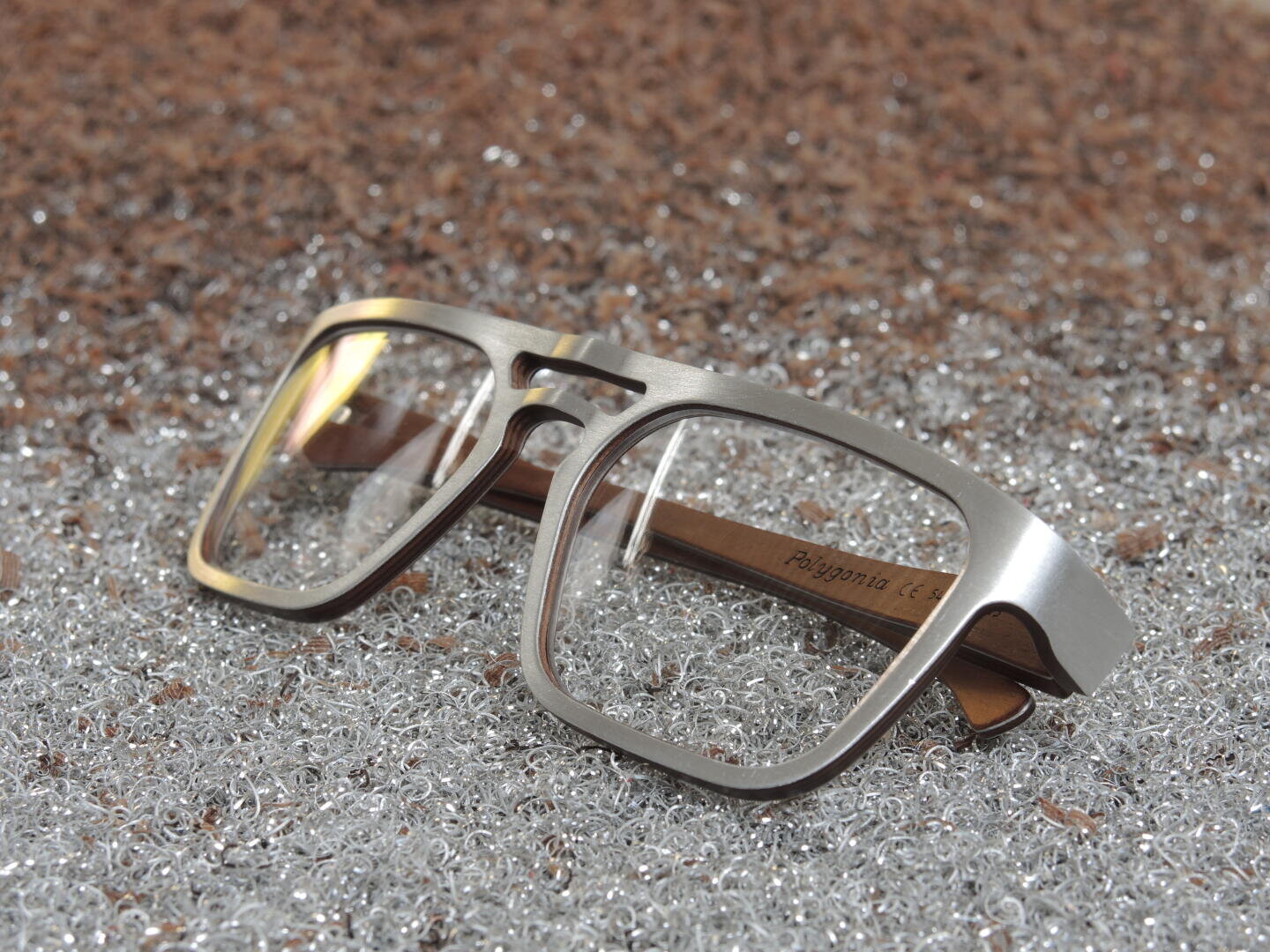 A pair of metal-rimmed eyeglasses with rectangular lenses and wooden arms rests on a sparkling, textured silver surface.