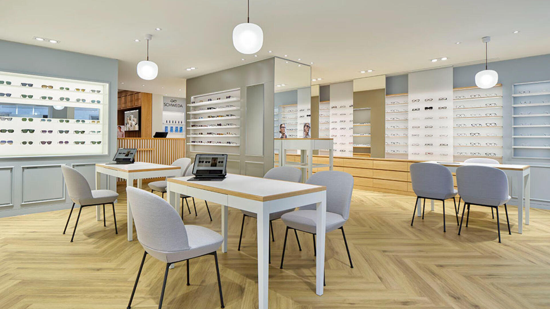Modernes Interieur eines Optikergeschäfts mit Holzboden, weißen Tischen und Stühlen, mit Brillen gefüllten Regalen an den Wänden und Laptops auf Tischen unter hängenden Pendelleuchten.