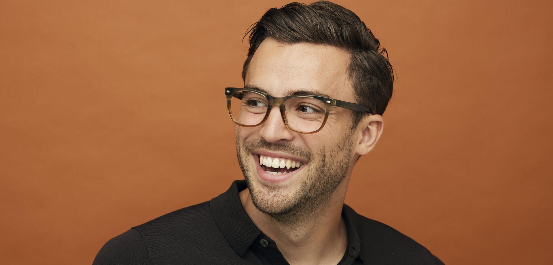 Ein Mann mit kurzen braunen Haaren, der eine Brille und ein schwarzes Hemd mit Kragen trägt, lächelt und blickt zur Seite vor einem einfarbig orangefarbenen Hintergrund.