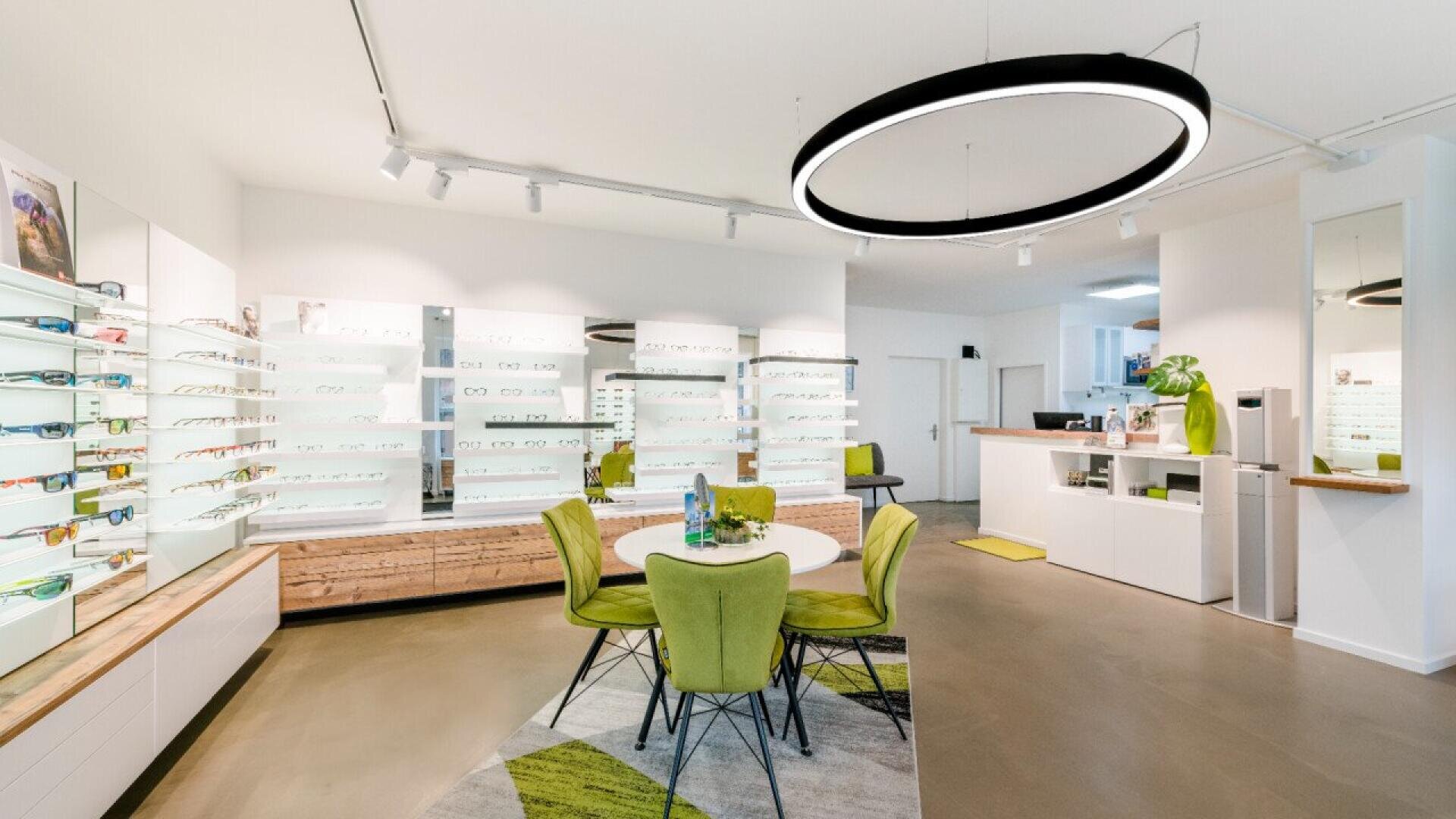 A modern optical store with bright white walls, green chairs around a round table, eyeglasses displayed on shelves, a large circular ceiling light, and decorative plants adding a fresh touch.