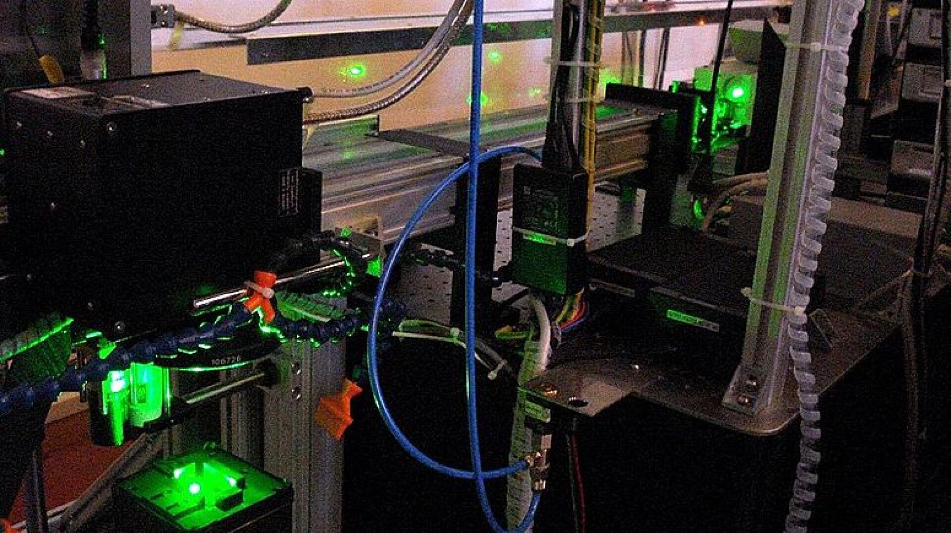 Laborgeräte mit Kabeln und Maschinen, die grünes Laserlicht ausstrahlen, verbunden durch Drähte und Schläuche, angeordnet auf einer Metallwerkbank in einem schwach beleuchteten Raum.