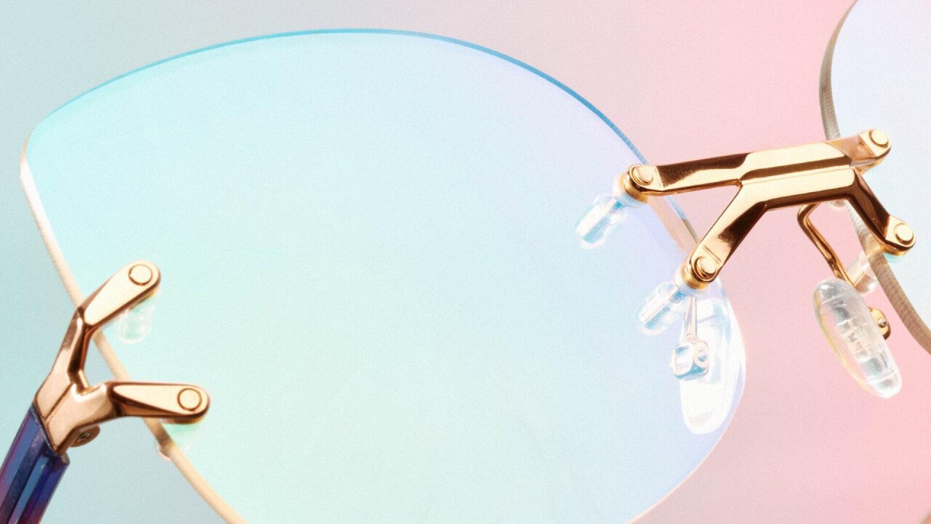 Close-up of stylish rimless eyeglasses with gold metal accents and nose pads, set against a soft pastel gradient background in shades of pink and blue.