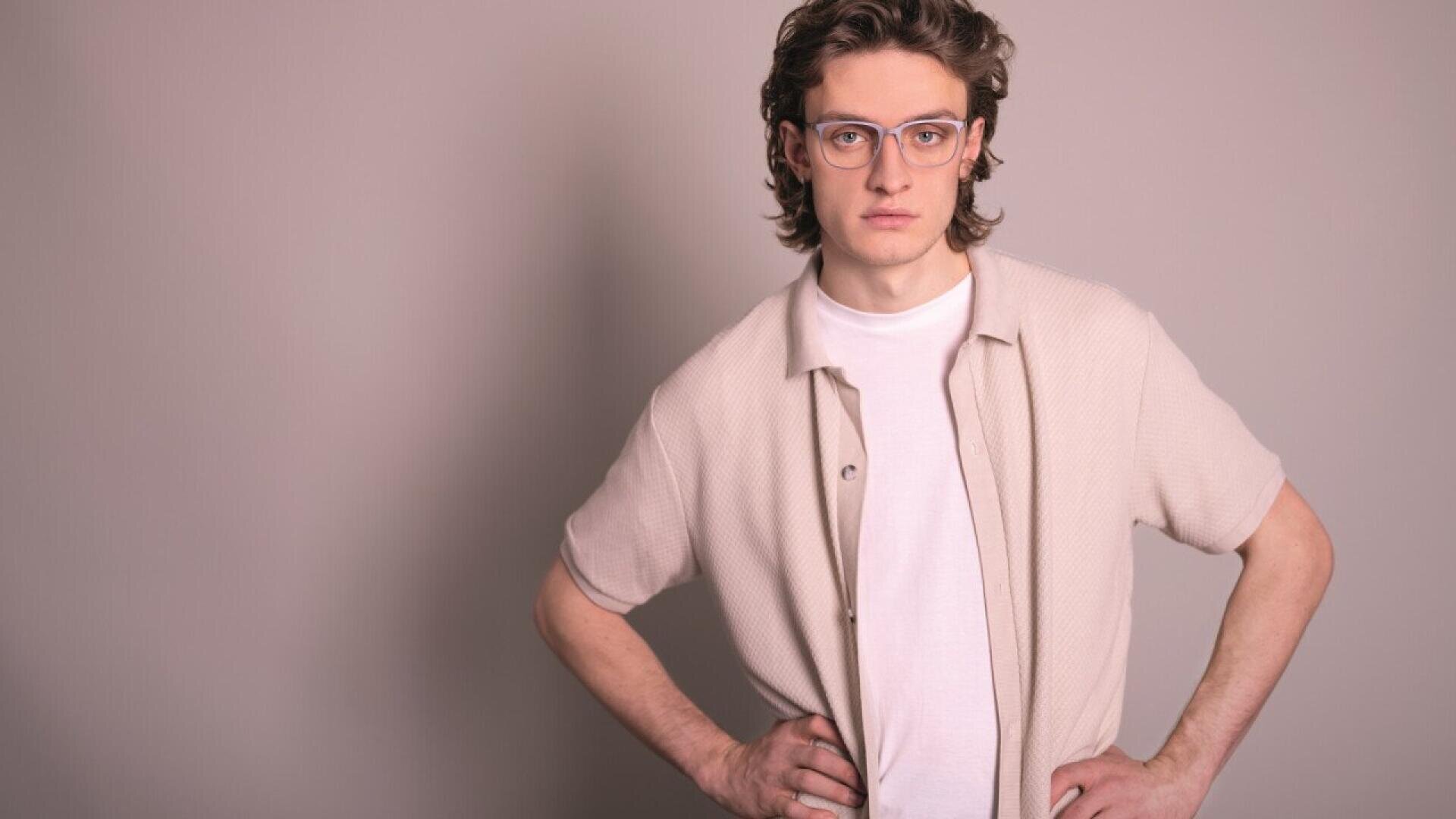 Young person with wavy brown hair and glasses stands with hands on hips, wearing a beige short-sleeve shirt over a white t-shirt, looking confidently at the camera against a plain light background.
